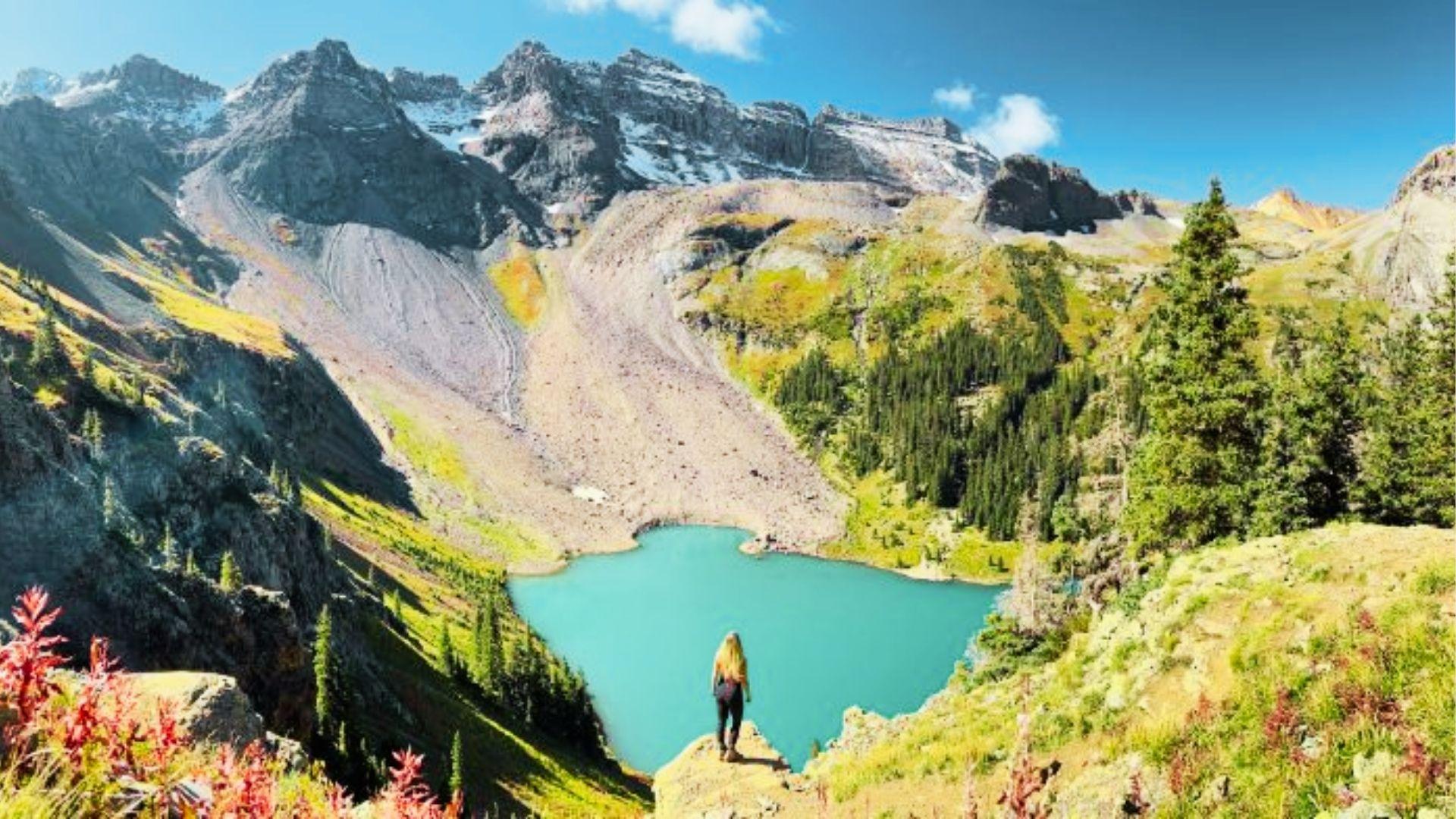 Woman with long hair stands on cliff overlooking turquoise alpine lake surrounded by rocky mountains