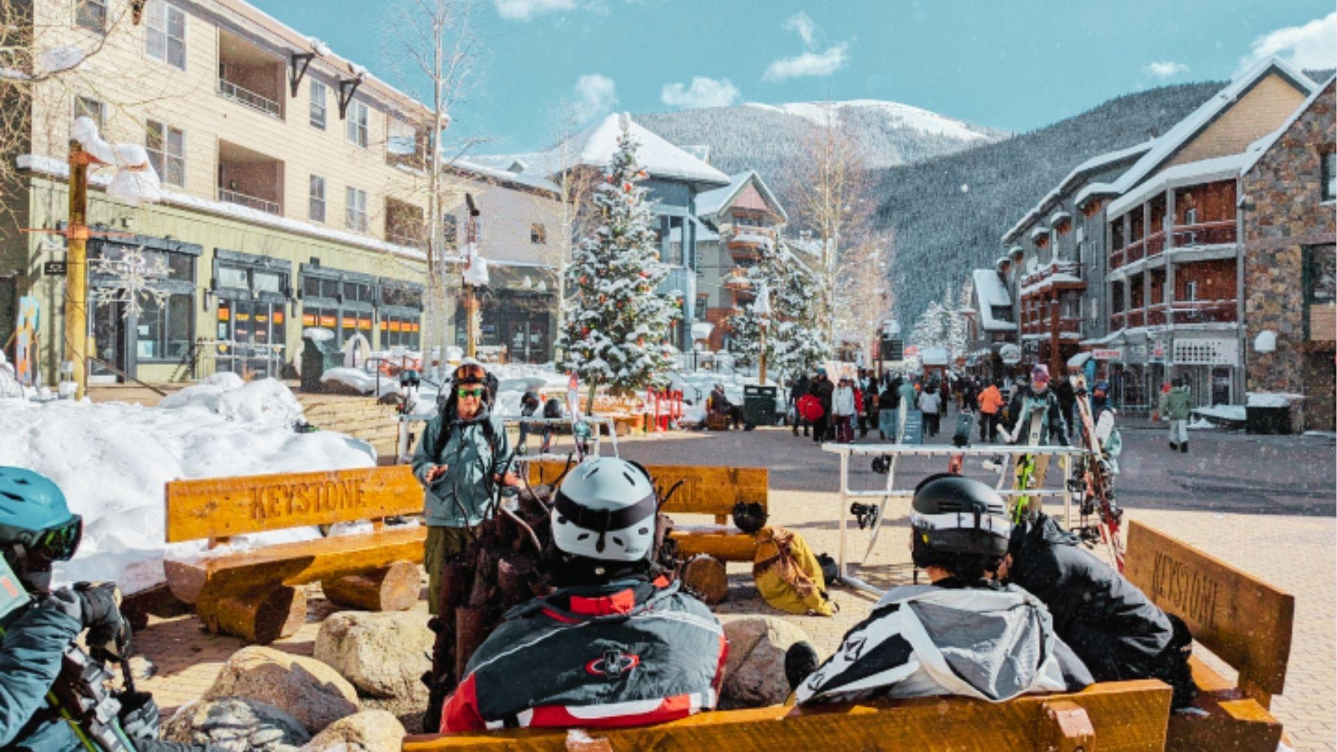 Skiers rest on Keystone benches in sunny village plaza with decorated tree and mountain backdrop