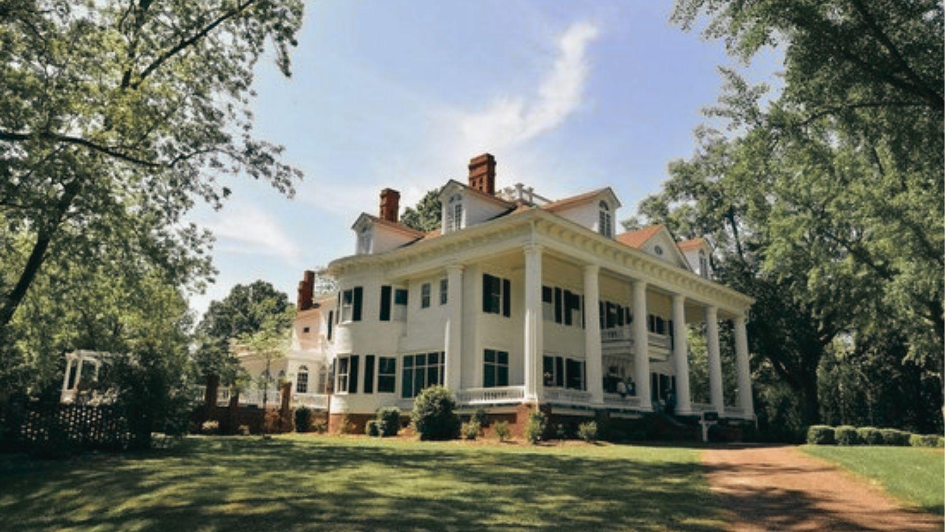 A stately white-columned mansion with a curved drive and mature trees under a blue sky.
