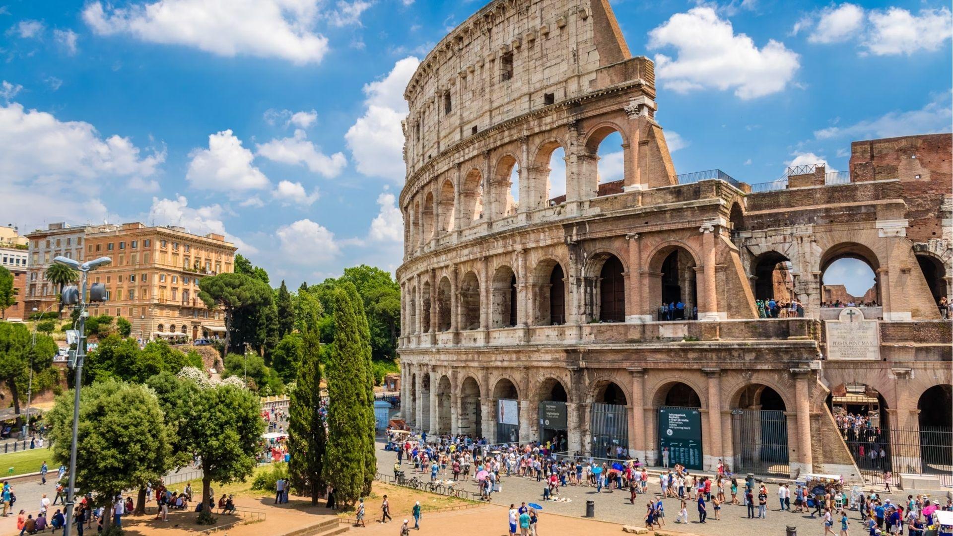 The ancient Colosseum stands under a bright blue sky in Rome, Italy, with tourists gathered at its entrance.