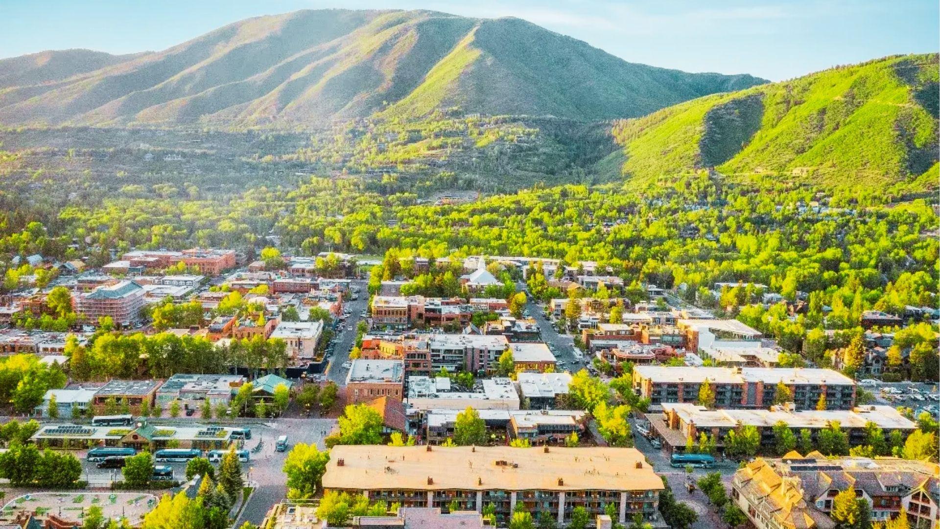 Sunlit mountain town aerial with hotels, buses, green trees, and ski slopes behind under blue
