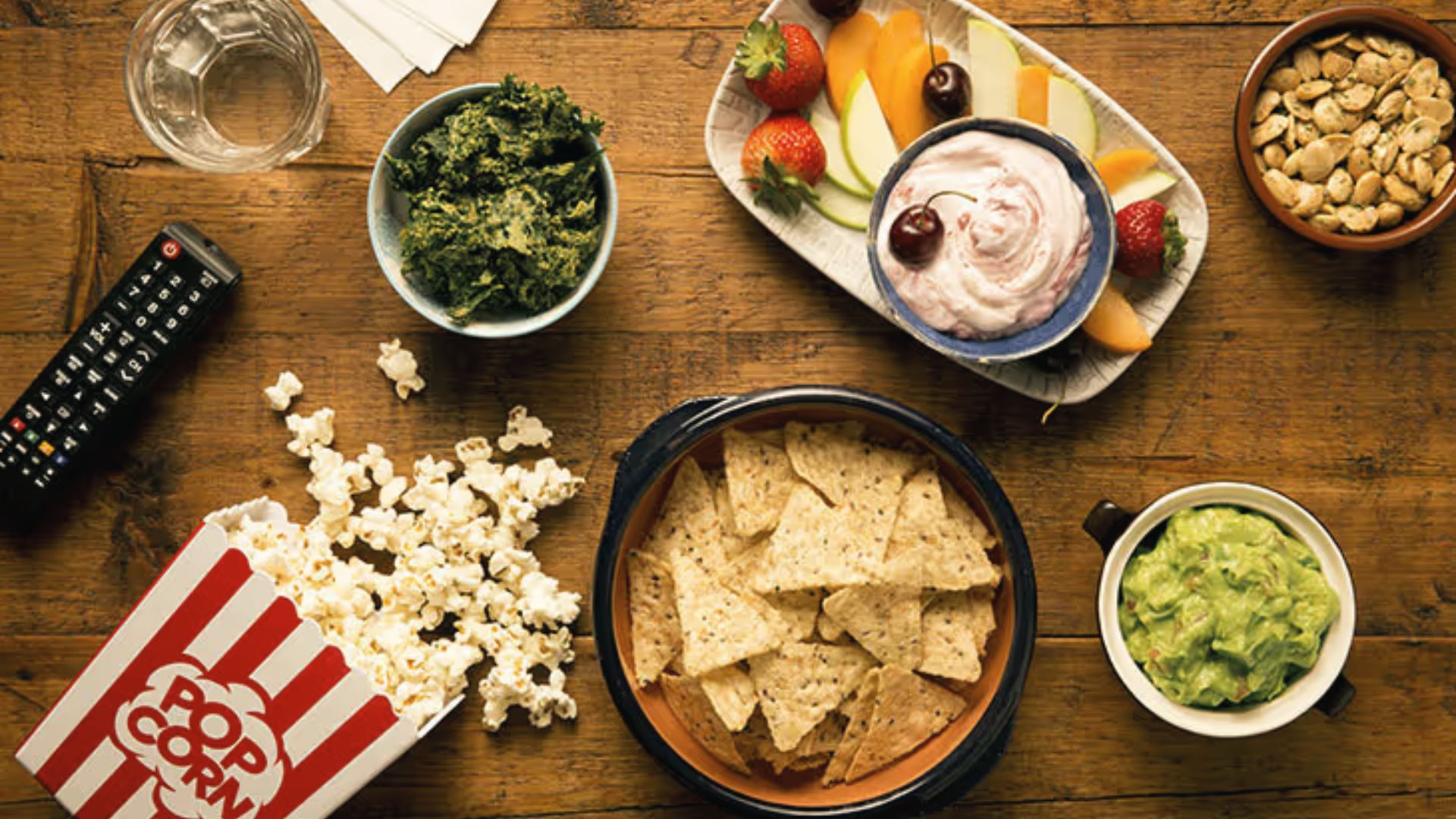 Flat lay of movie night snacks on a rustic wood table, including popcorn, tortilla chips with guacamole, kale chips, and fresh fruit.