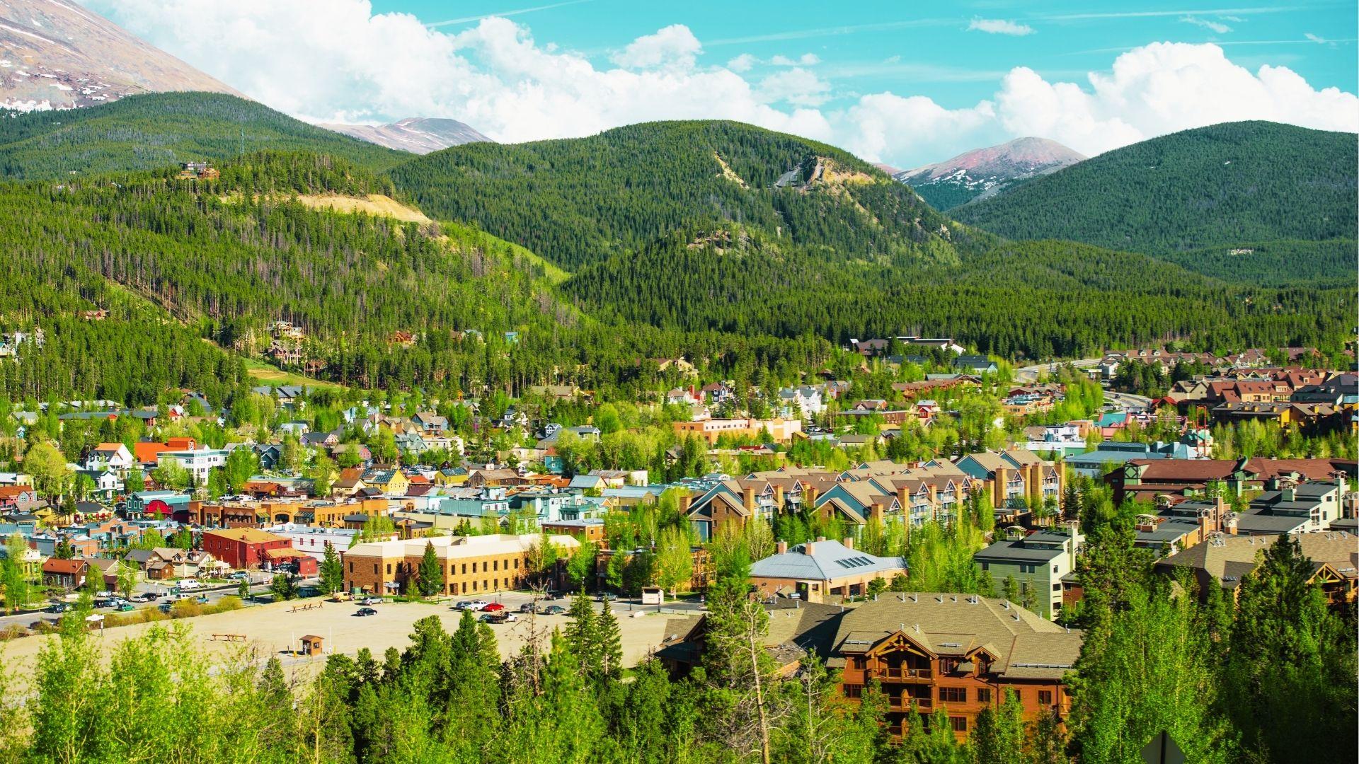 Aerial of mountain town with colorful buildings, green trees, ski lodges, and forested hills surrounding