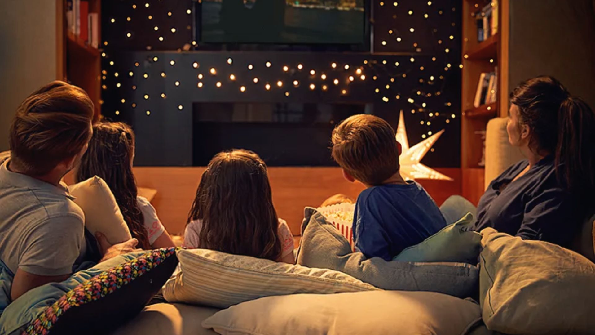 A family of five watches TV from a couch surrounded by warm string lights and a glowing star lantern.