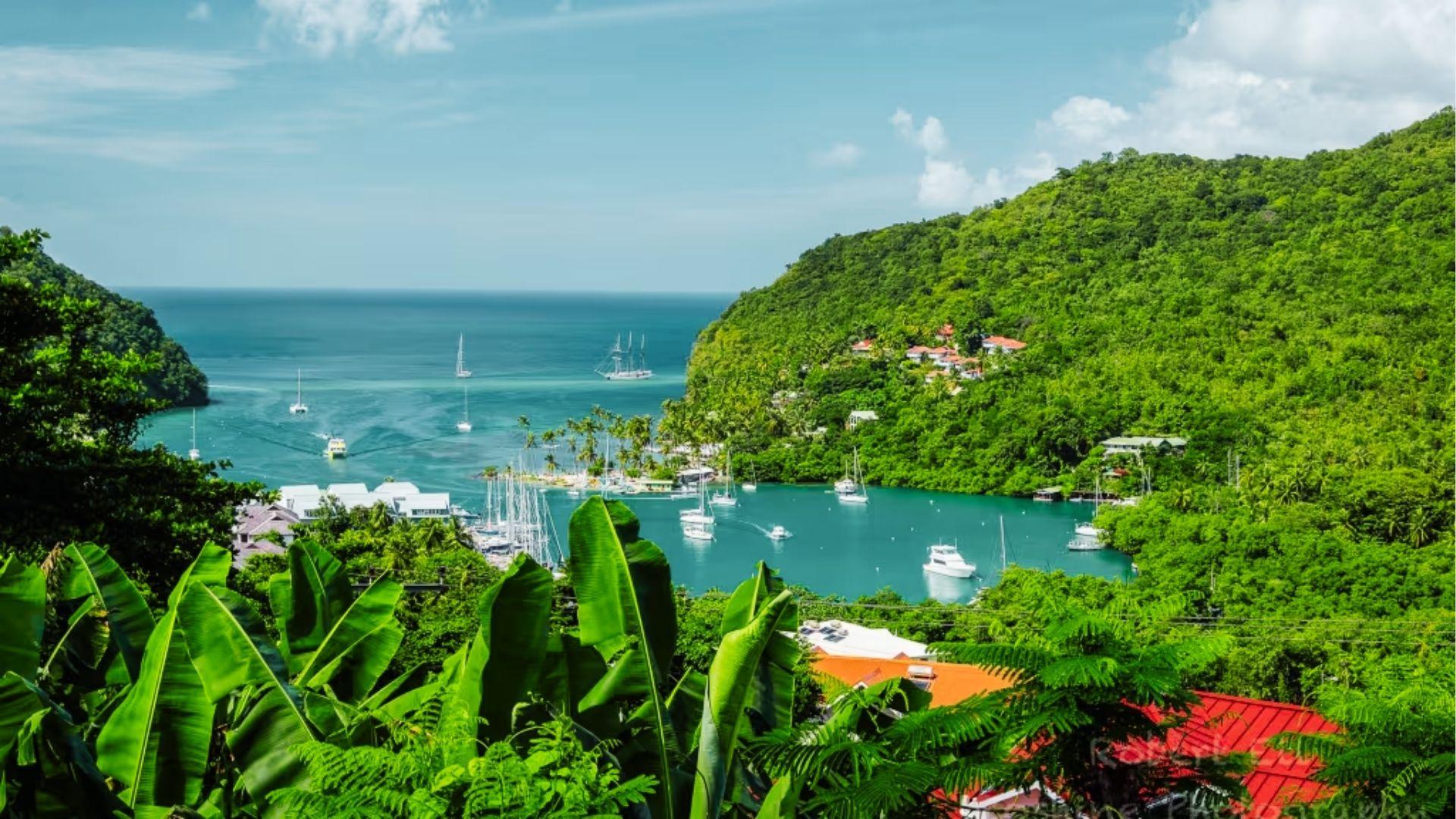 Lush green hills frame turquoise bay with sailboats, marina, and village nestled by ocean