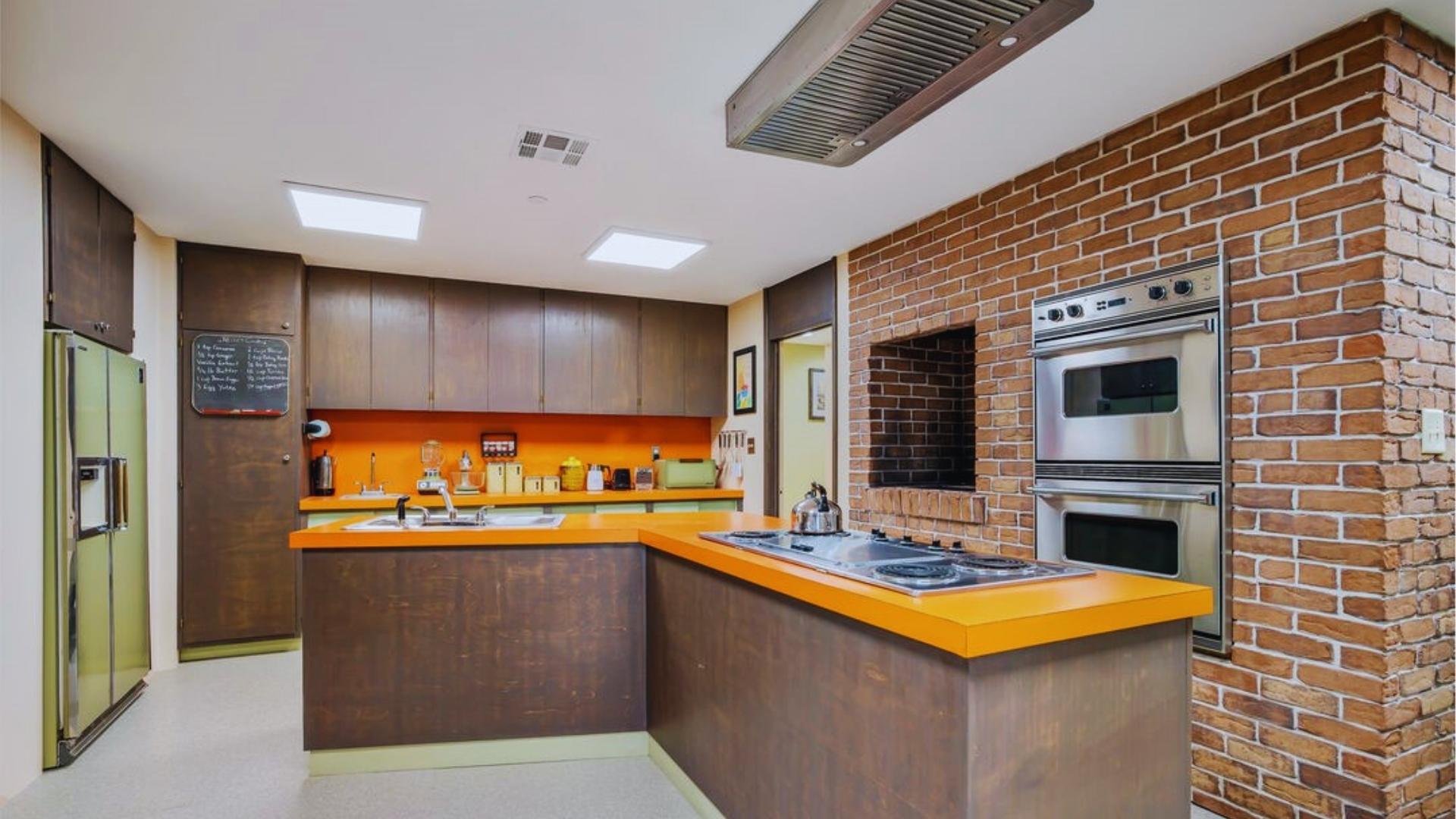 Brady Bunch kitchen with orange Formica counters, brick wall, double ovens, and avocado green fridge.