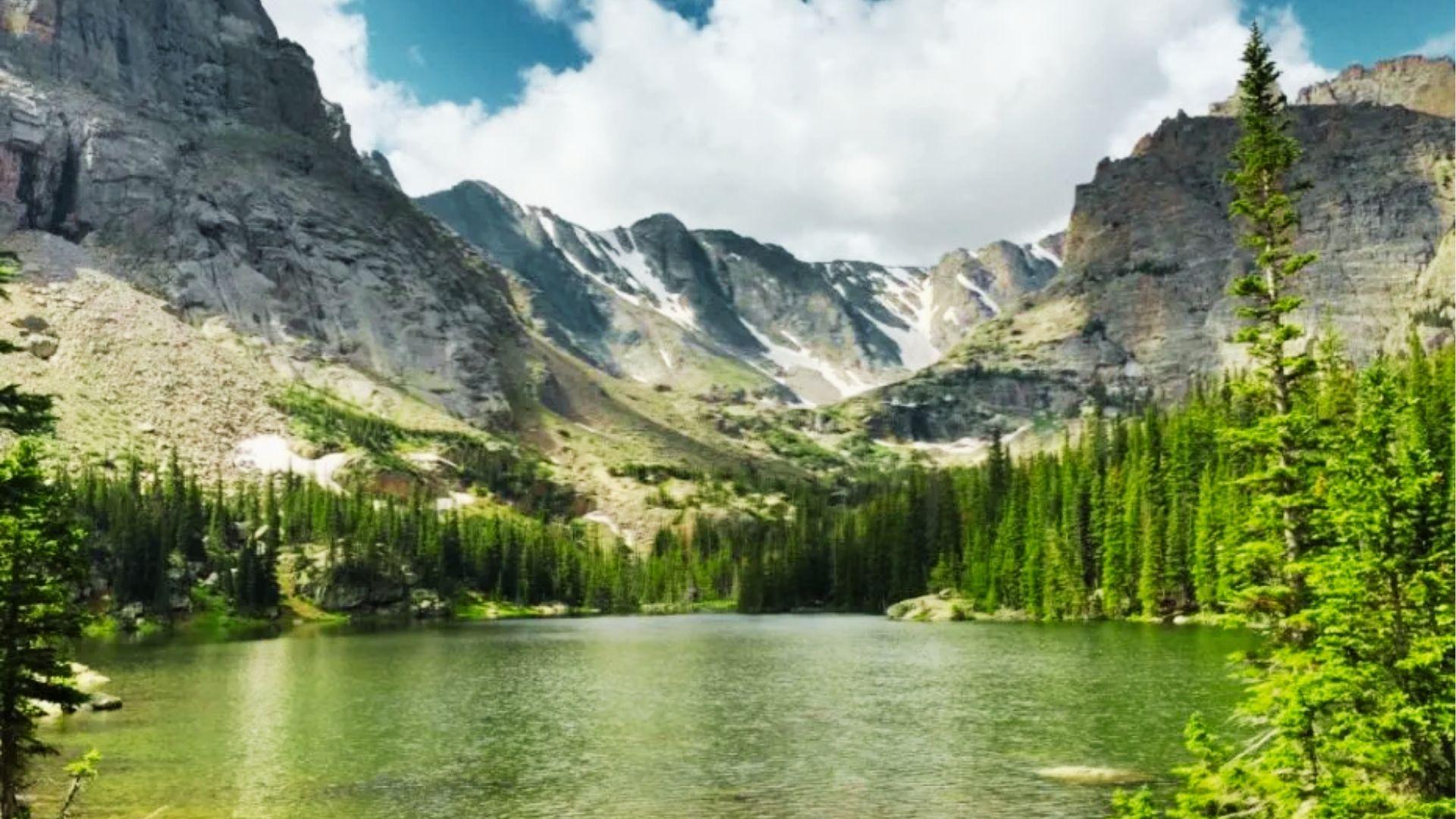 Calm alpine lake surrounded by pine forest and rugged snowcapped mountains under partly cloudy sky