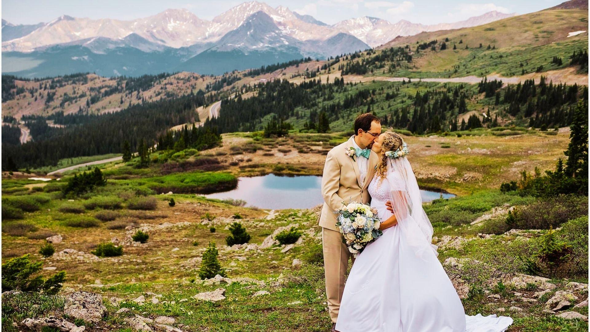 Bride and groom kiss on mountain ridge overlooking alpine lake, forest, and snowcapped peaks