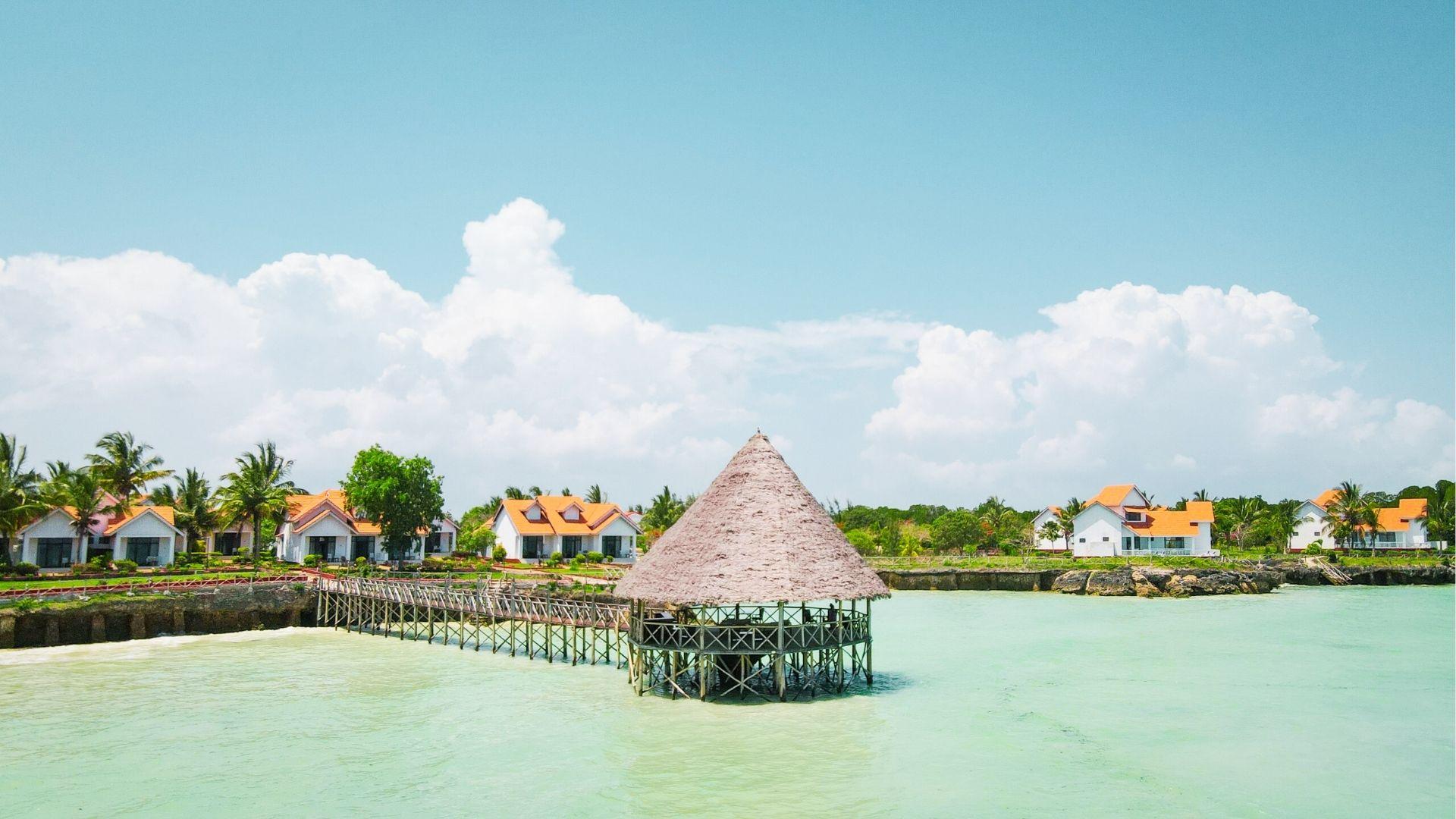 Thatched roof hut on stilts over turquoise water with wooden pier, resort villas, palms, clouds