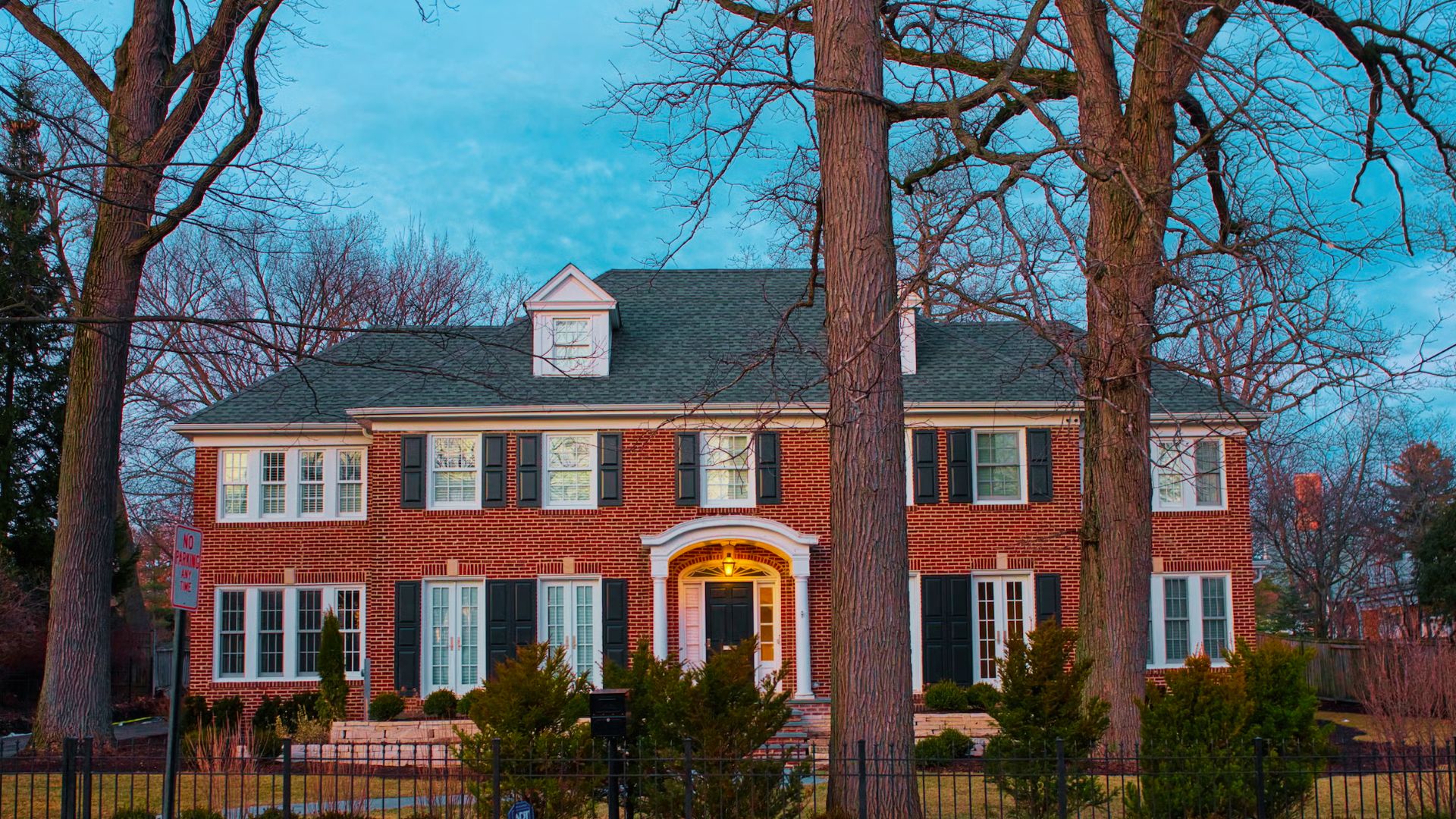 A grand red-brick Georgian home with black shutters and a lit entryway, framed by tall winter trees.