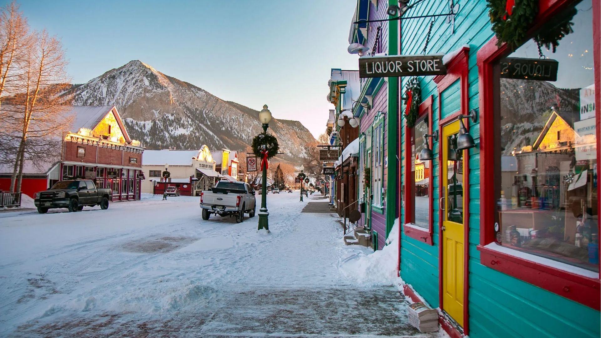 Snowy main street with colorful shops, liquor store, parked trucks, mountains, and holiday wreaths