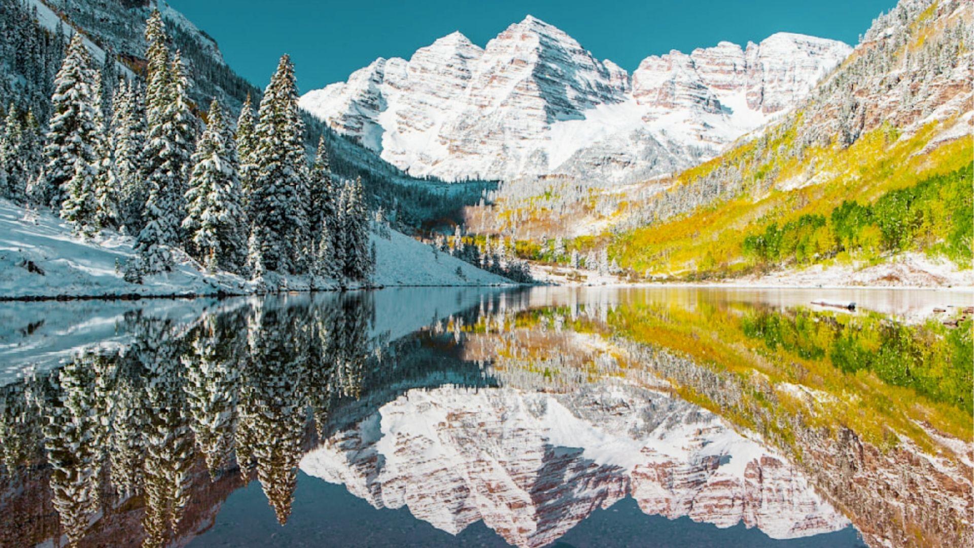 Snow covered Maroon Bells peaks reflect in lake with evergreen trees and autumn hillsides glowing