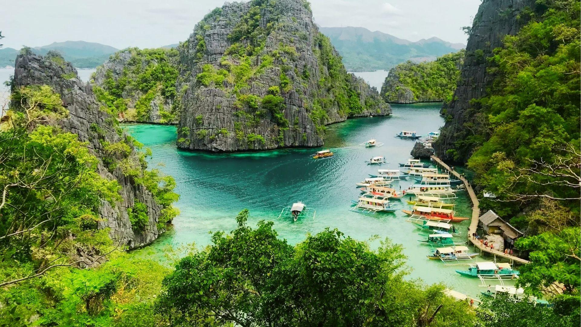 Limestone cliffs surround emerald lagoon with docked boats and wooden walkway through lush forest