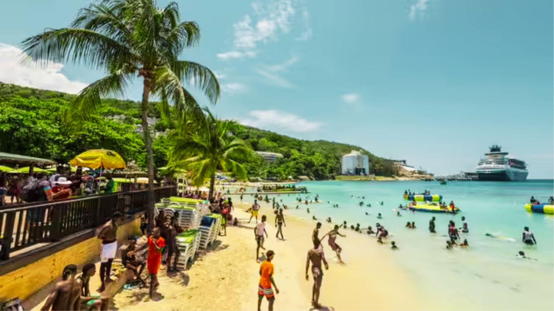 Crowded sandy beach with swimmers, palm trees, inflatable toys, and cruise ship offshore