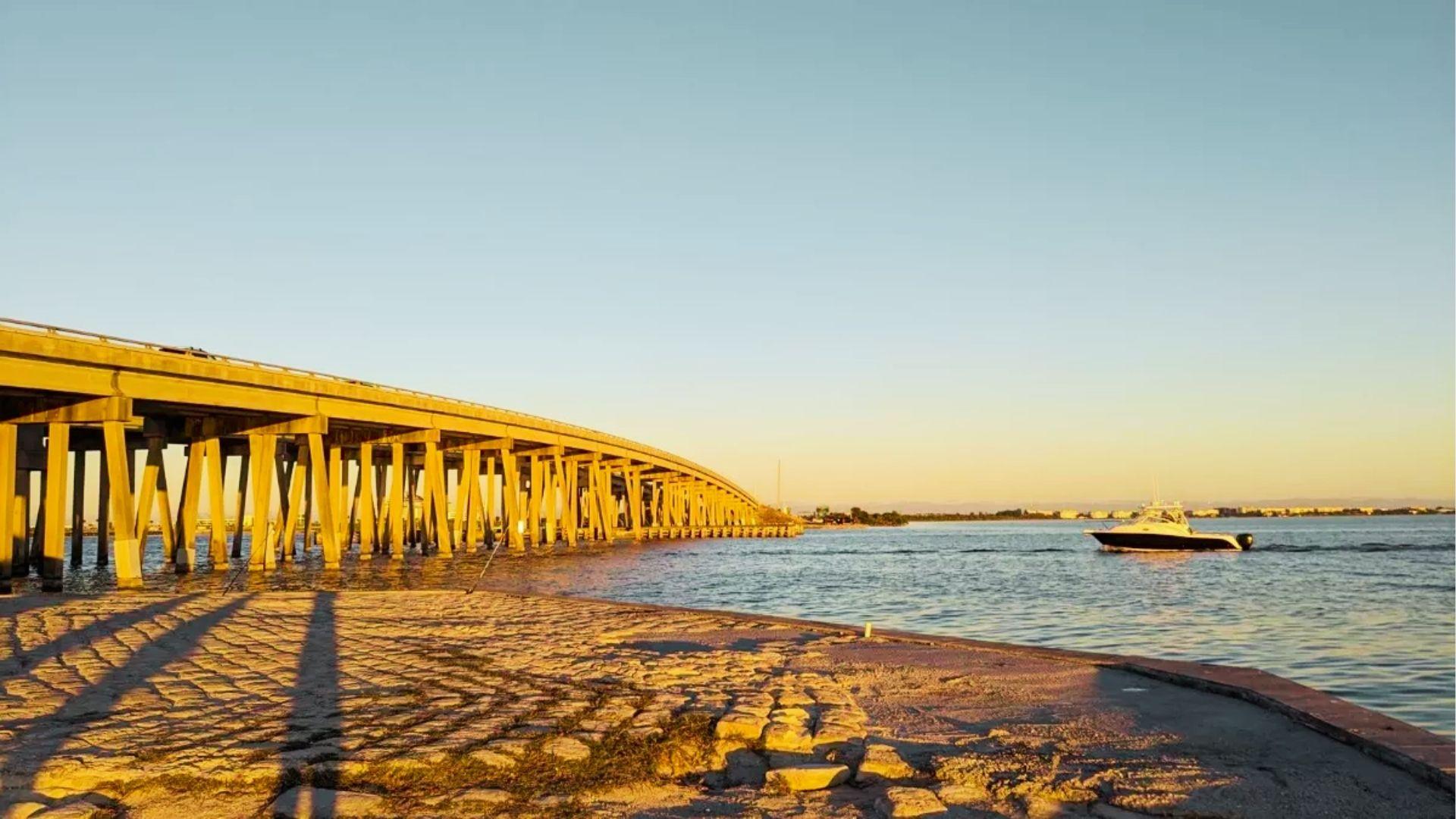 Boat cruises past concrete bridge at golden hour with long shadows on rocky shoreline