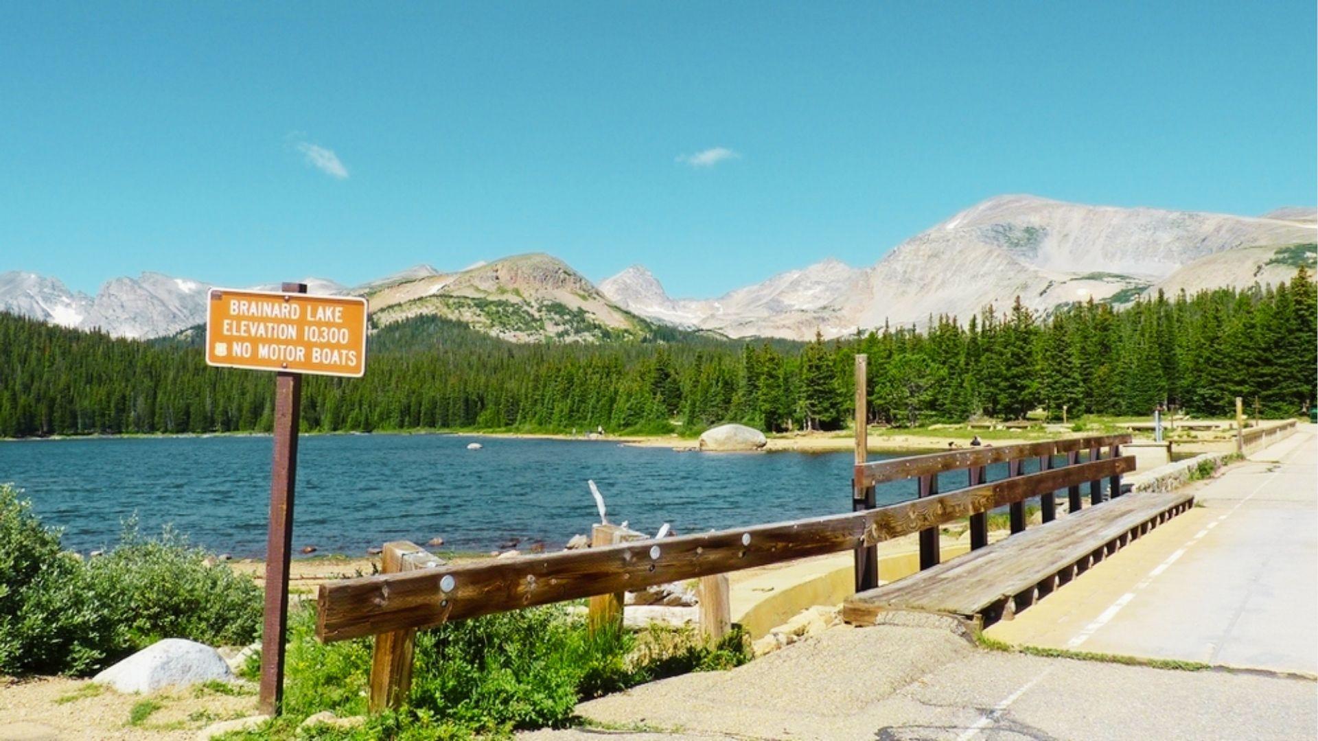 Brainard Lake sign at 10,300 feet elevation stands by bridge with pine forest and peaks