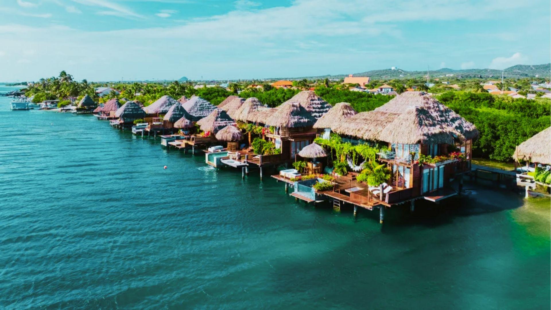 Thatched roof overwater bungalows with private decks line clear blue lagoon beside lush coastline