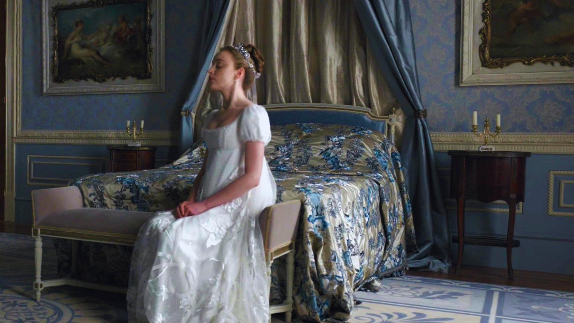 Daphne Bridgerton in white empire-waist gown seated in blue silk bedroom with floral bedding and canopy.
