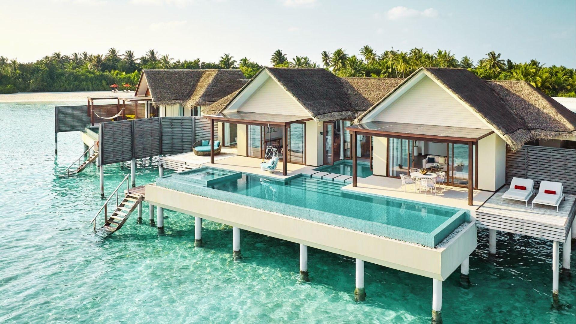 Luxury overwater villas with private pools extend above clear turquoise water near palm lined shore