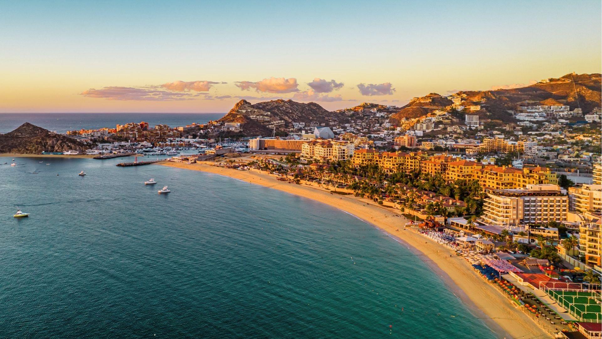 Golden sunset hits the curved beach of Cabo San Lucas, with luxury resorts and boats dotting the bay.