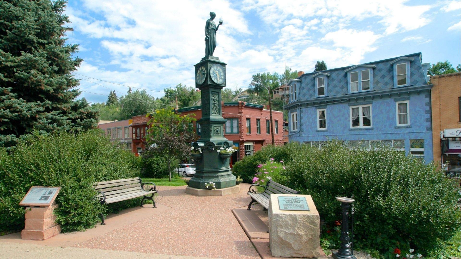 Historic town square with clock tower statue, benches, flowers, and colorful buildings on summer day