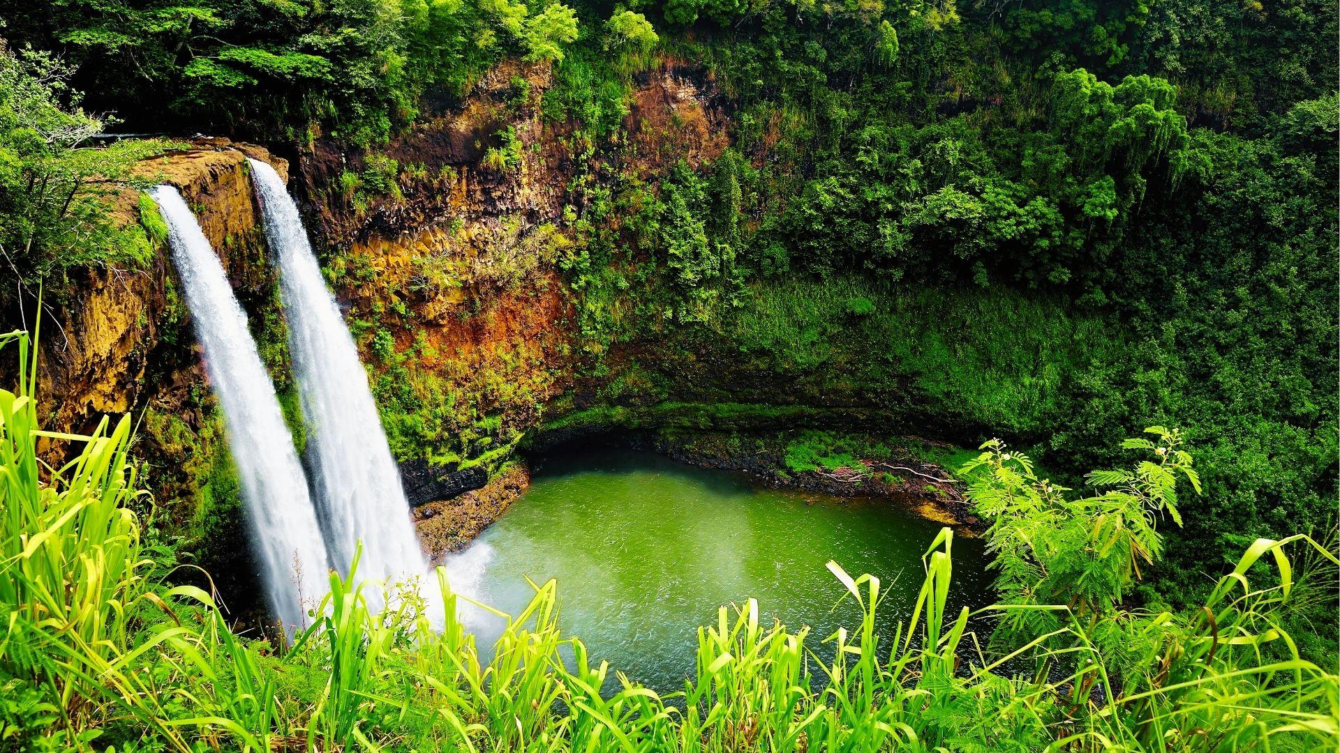 Twin waterfalls cascade into emerald pool surrounded by dense green tropical forest and cliffs