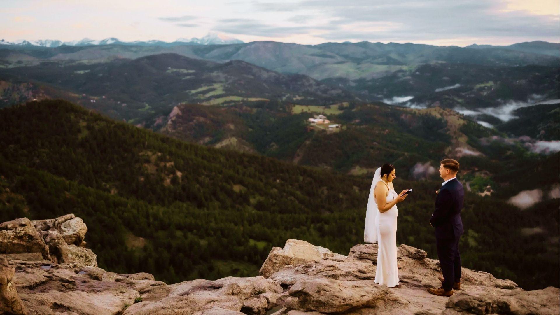 Bride reads vows to groom on rocky mountain cliff overlooking misty forested valleys and snowcapped peaks