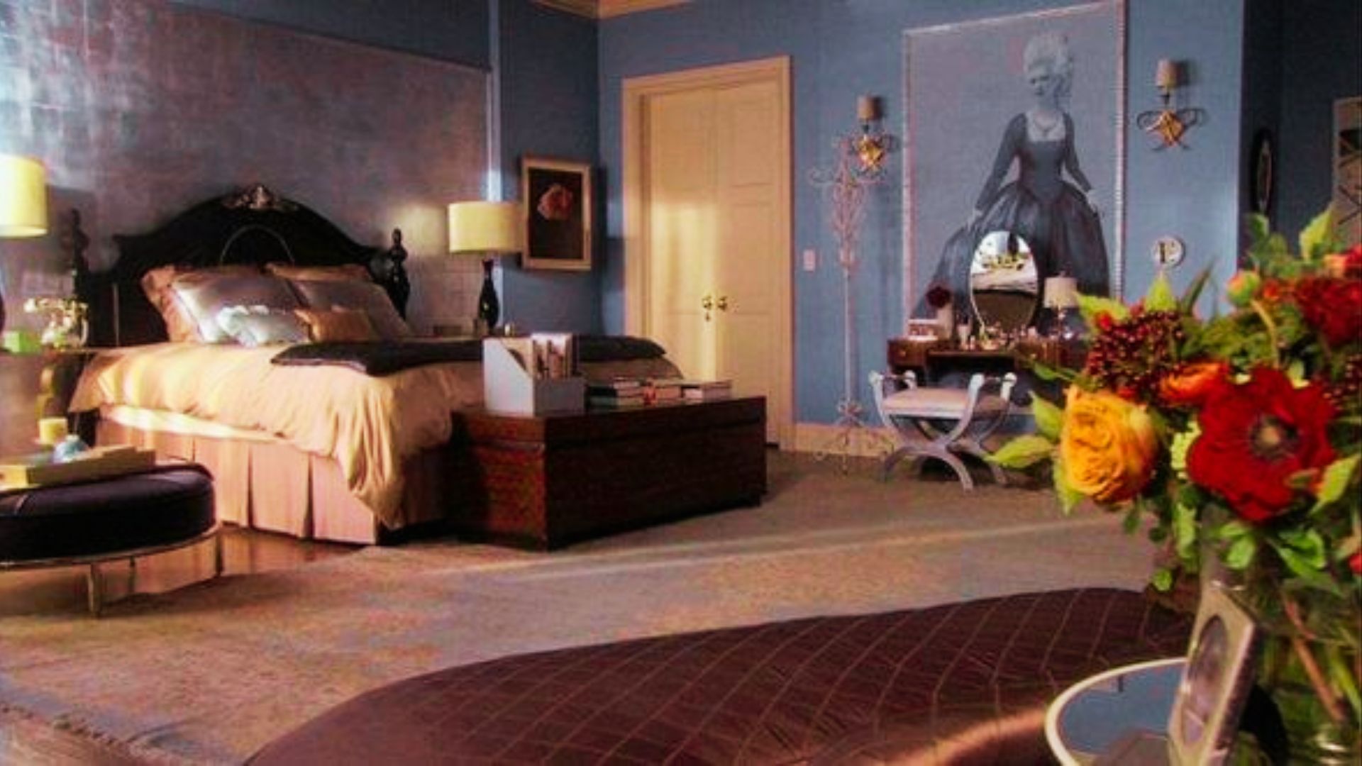Wide view of Blair Waldorf’s blue bedroom featuring a dark wood bed with gold bedding, silver-leaf accent wall, and twin table lamps.