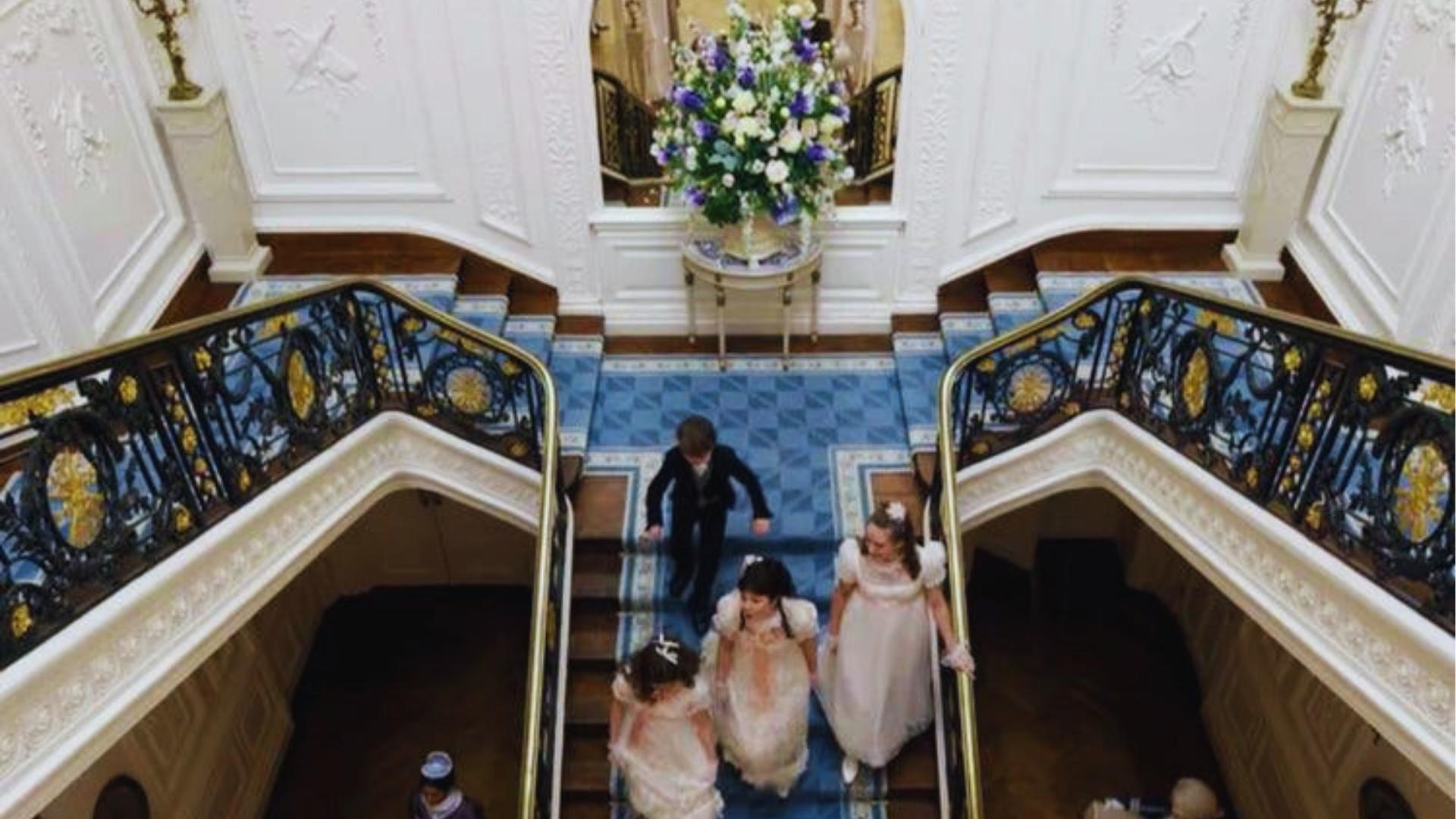 Grand double staircase in Bridgerton house with blue carpet runner, wrought iron railings, and floral arrangement.