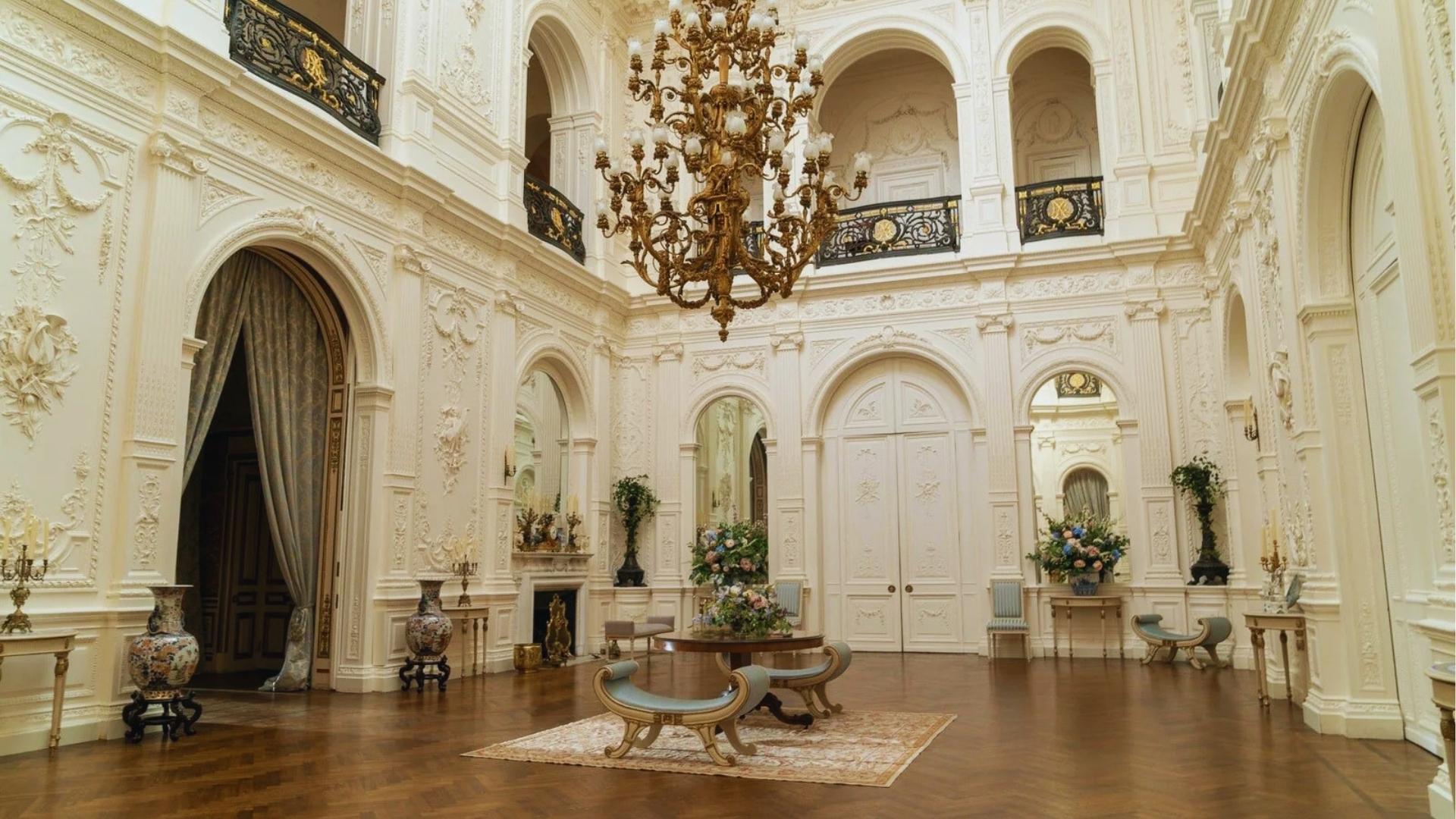Bridgerton ballroom entrance hall with white plasterwork, massive gilded chandelier, and Chinese porcelain vases.
