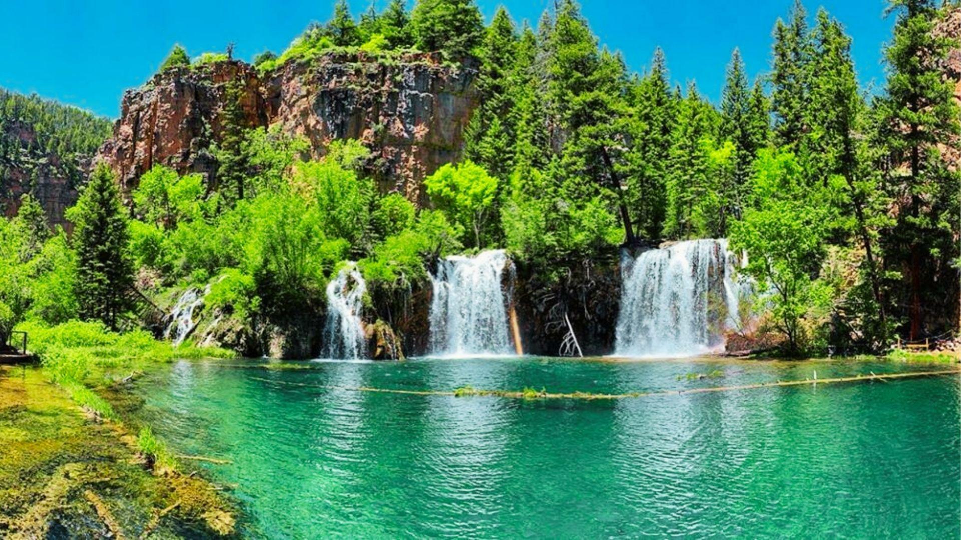 Multiple waterfalls cascade over mossy cliffs into turquoise pool surrounded by lush green forest