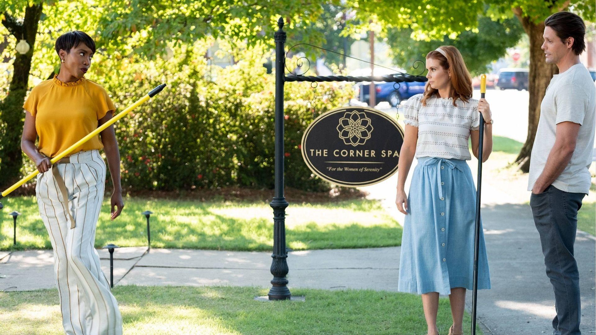 Three actors stand outside “The Corner Spa” sign, holding cleaning tools on a sunny lawn.