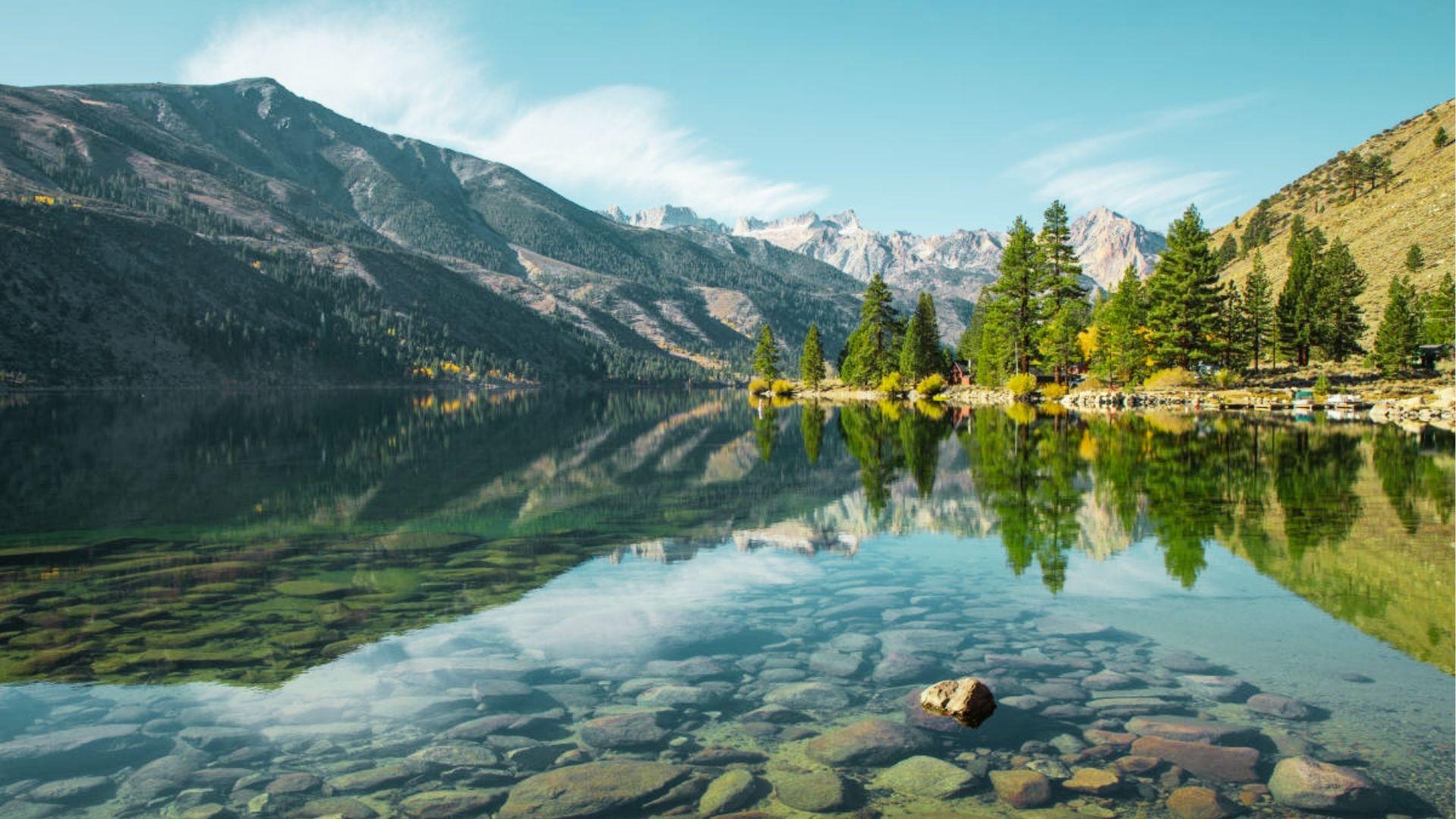 Crystal clear alpine lake reflects mountains, pine trees, rocks, and clouds on calm summer day