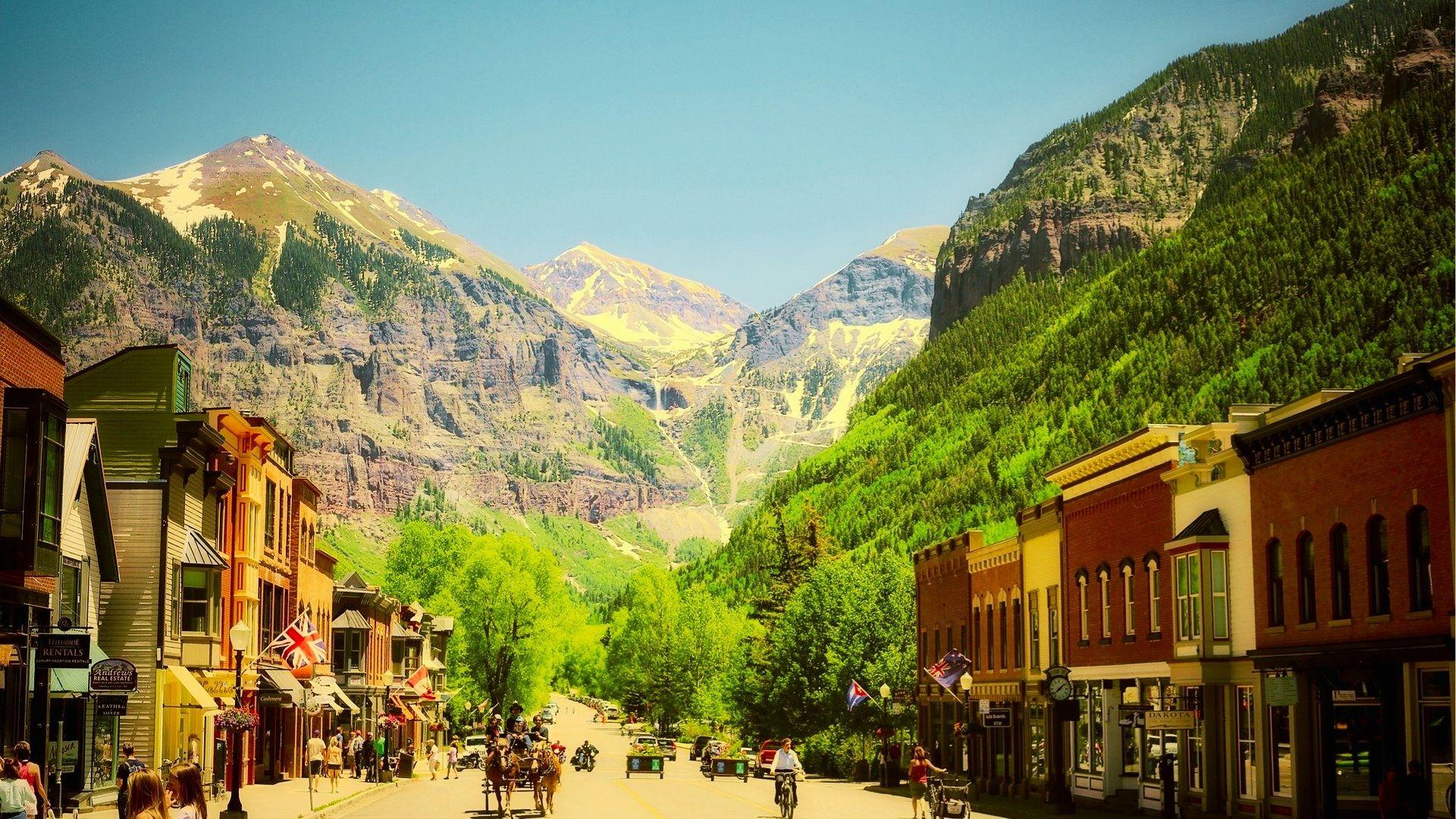 Sunny mountain town street lined with shops, flags, horse carriage, cyclists, and forested peaks beyond