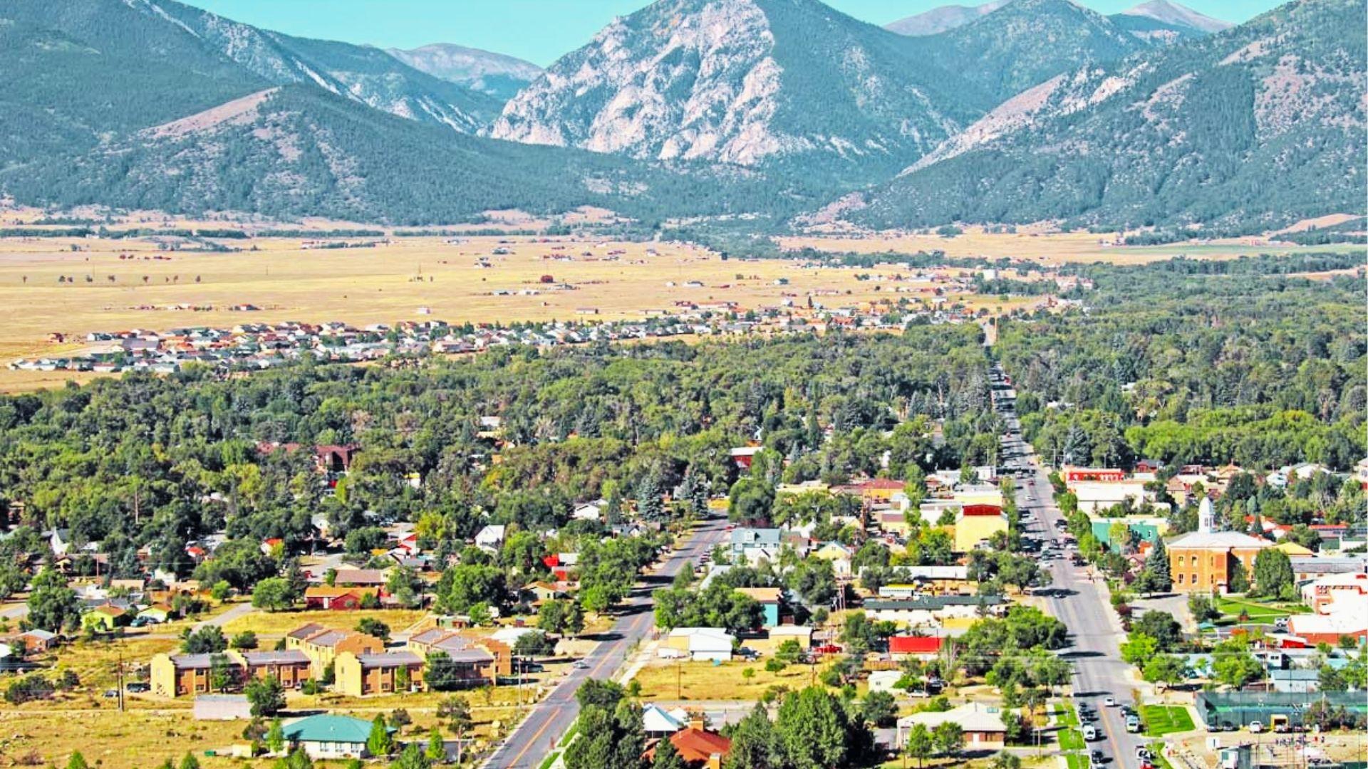 Aerial view of mountain town streets, homes, and forests with rugged peaks under blue sky