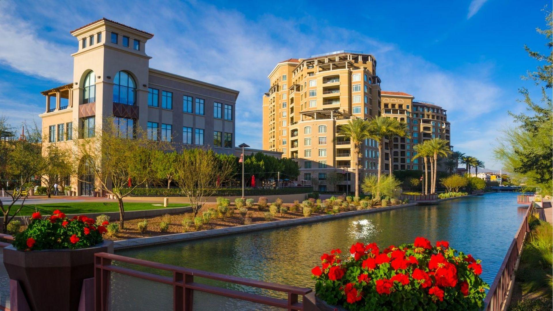 Modern resorts with palm trees and red flowers border a canal under bright blue skies in a desert city.