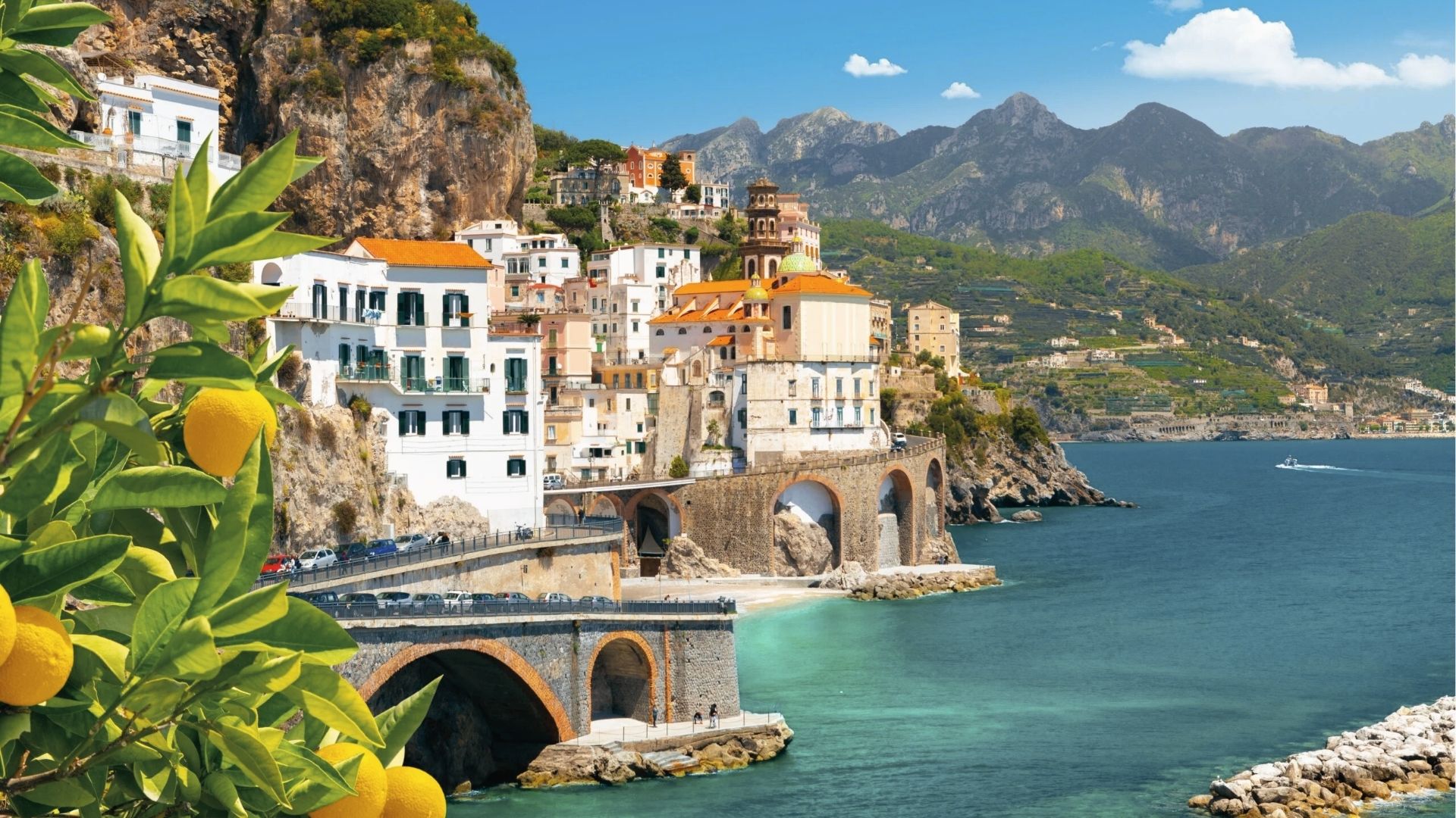 Bright Amalfi lemons hang from a tree in the foreground as the town of Atrani clings to rocky cliffs.