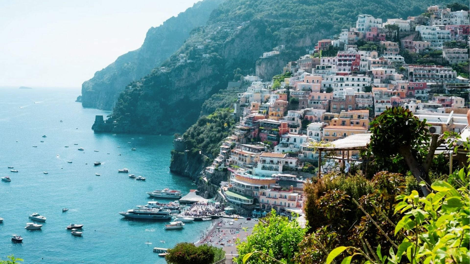 Colorful cliffside buildings stack above the harbor in Positano, with turquoise water dotted by boats. 
