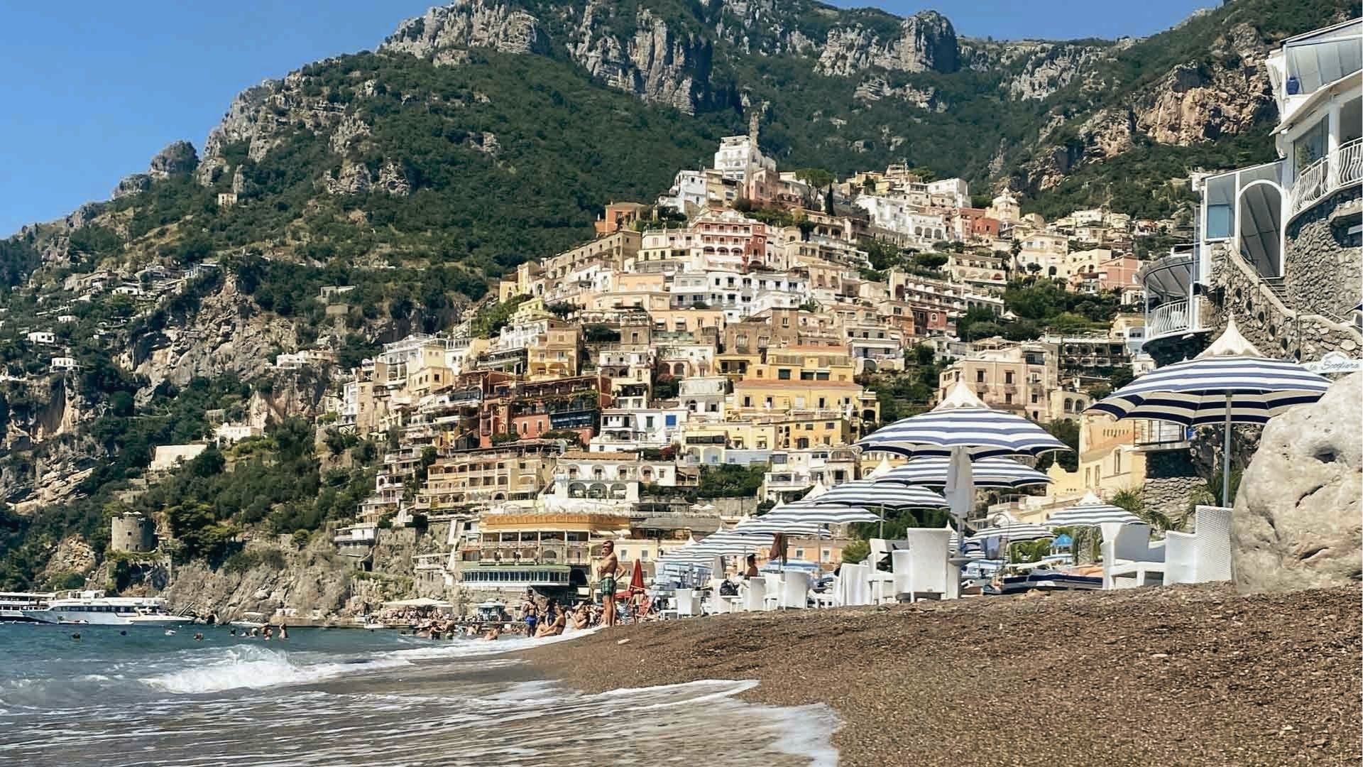 Striped blue and white umbrellas shade a pebbled beach in Positano as waves meet the shore. 
