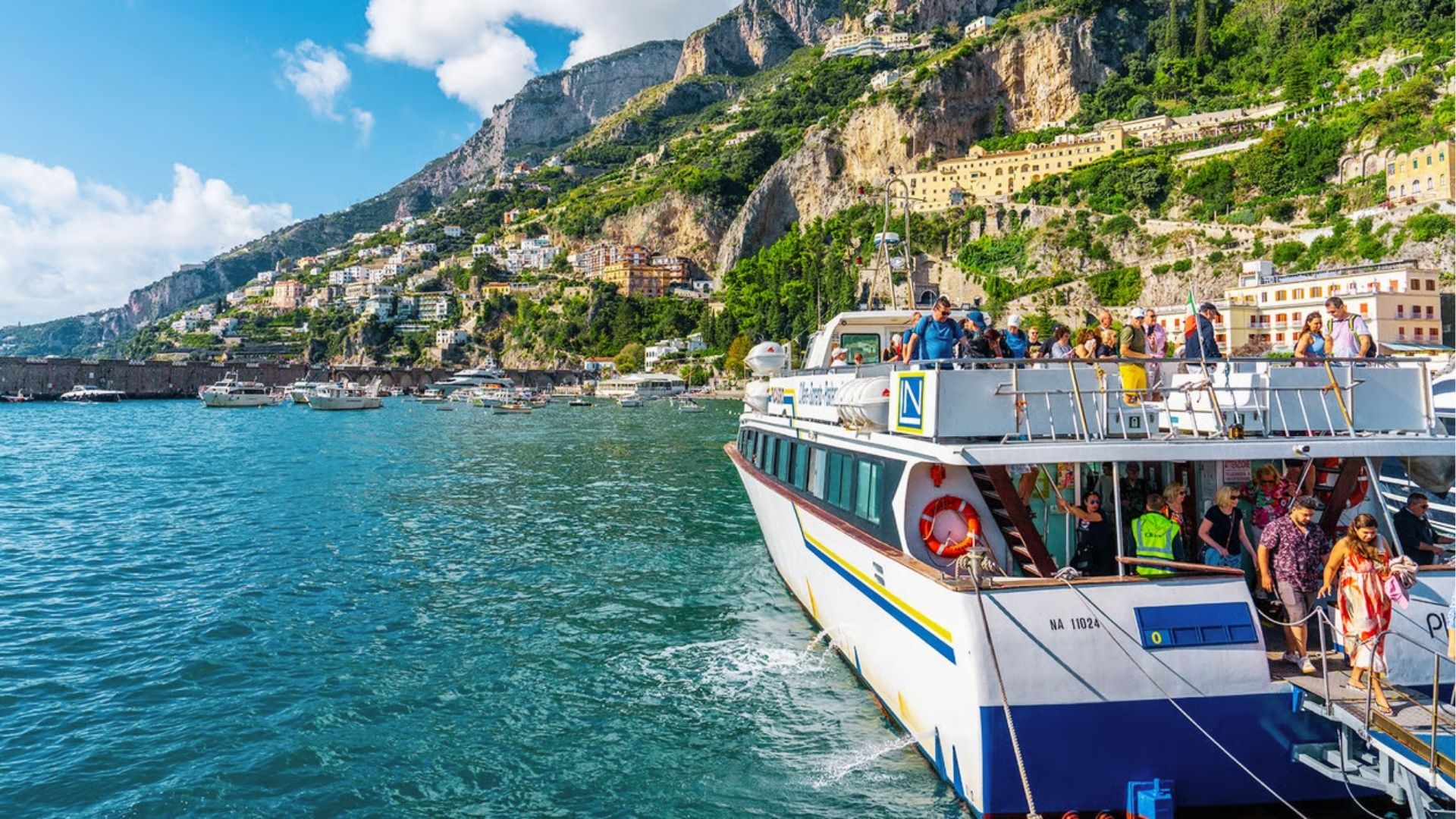 Tourists disembark a ferry at the port, with the Amalfi Coast’s green cliffs and stacked villages behind them. 
