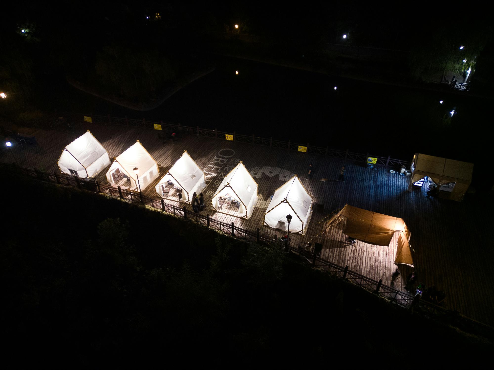 A cluster of tents illuminated by soft lights on a dock, surrounded by calm water under a night sky.