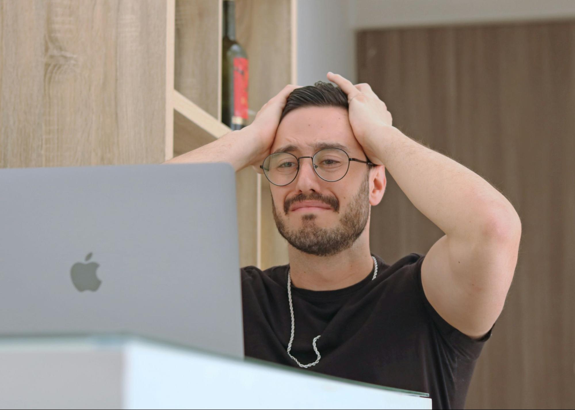 A man wearing glasses rests his head on his hands while sitting at a desk, appearing deep in thought or contemplation.