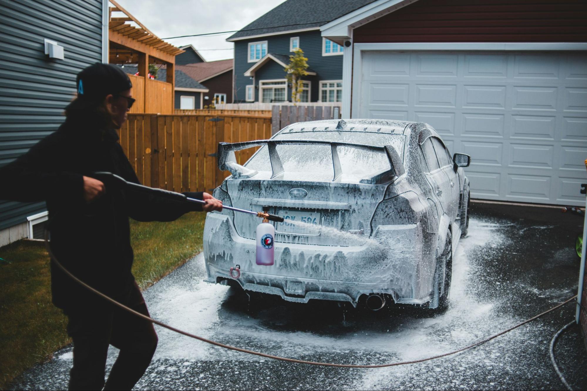A woman using a pressure washer to clean her car in a driveway, surrounded by greenery.