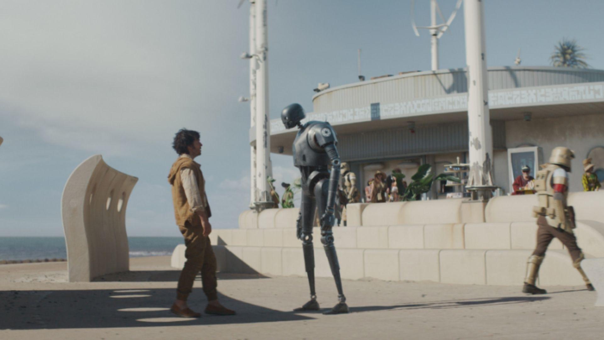 Cassian Andor faces droid K-2SO on a sunny beachfront promenade with alien extras nearby.