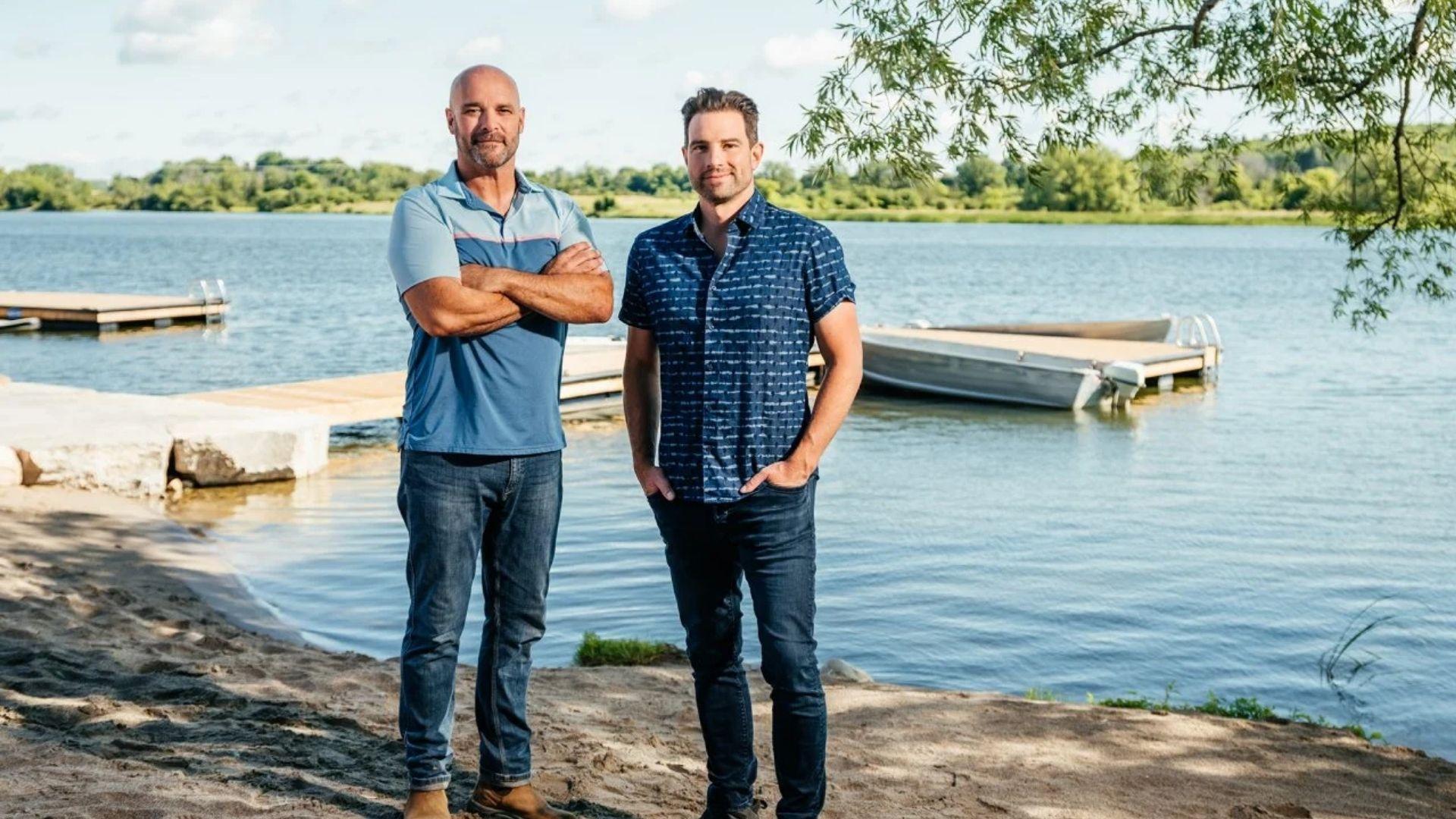 Renovation Resort Showdown. Two male hosts pose on a sandy lakeside shore with a dock.