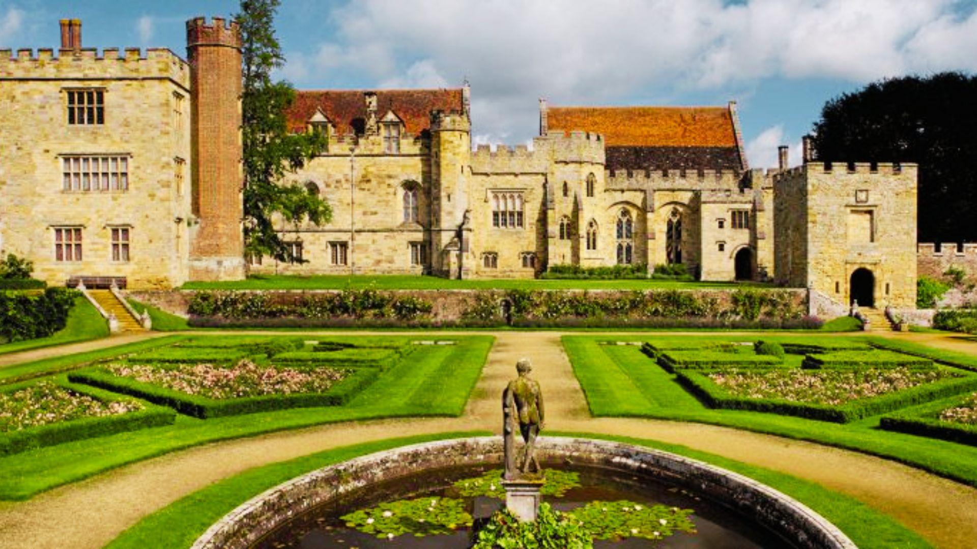 Sunlit formal gardens with a central fountain stretch before Penshurst Place, a medieval manor in Kent.