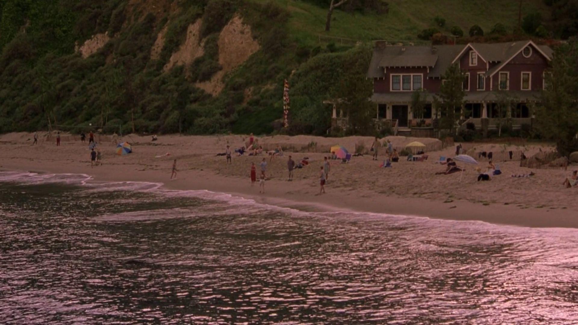 A busy beach scene with sunbathers and swimmers below a green cliff and red beach house.