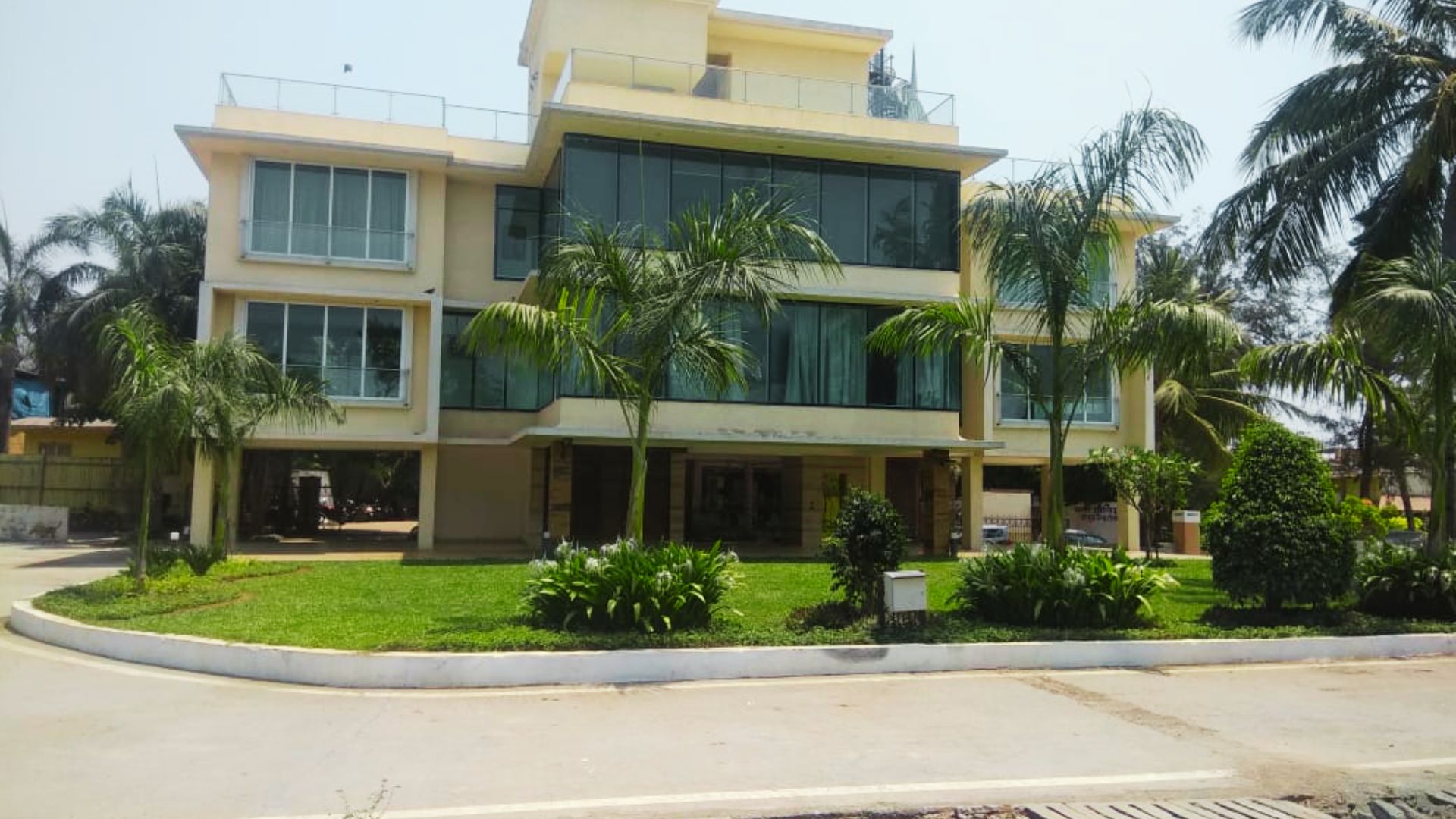 Large modern 3-story beige building with glass walls and palm trees, surrounded by a circular driveway.