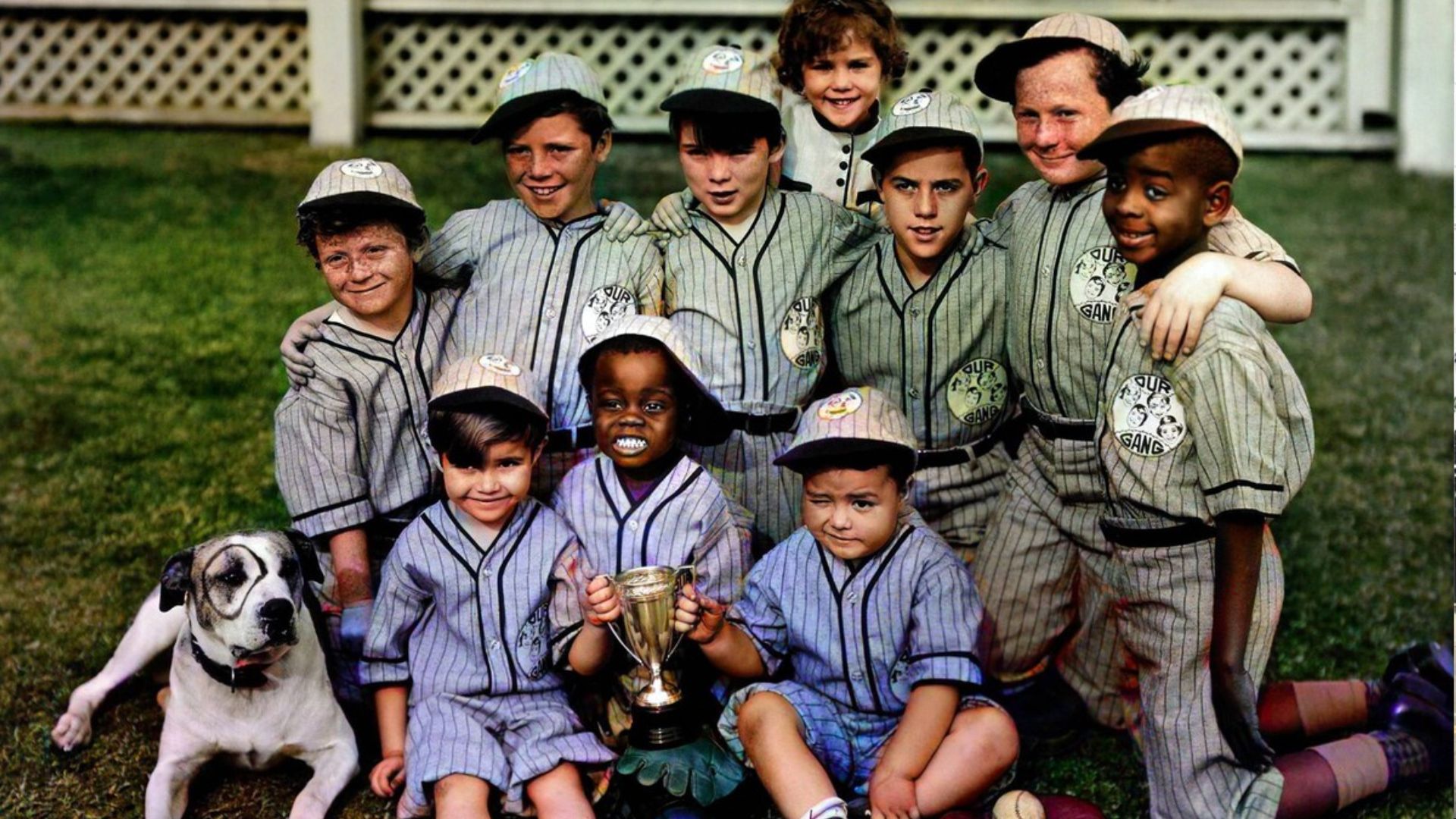 Original Our Gang cast in matching baseball uniforms posing with a trophy and their dog Pete on a grass lawn in a vintage photo