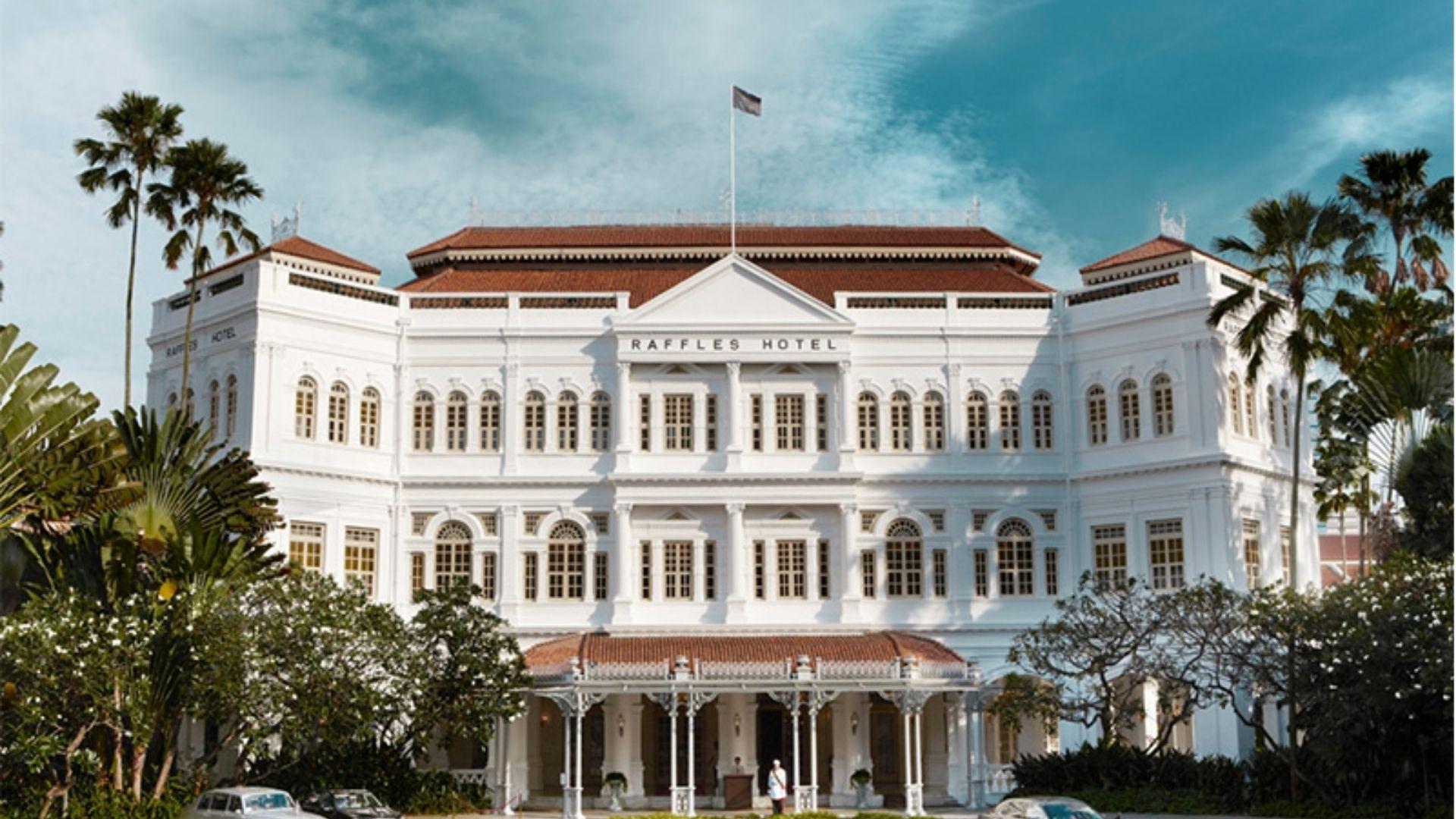 The white colonial facade of Raffles Hotel Singapore flanked by palm trees under a blue sky.